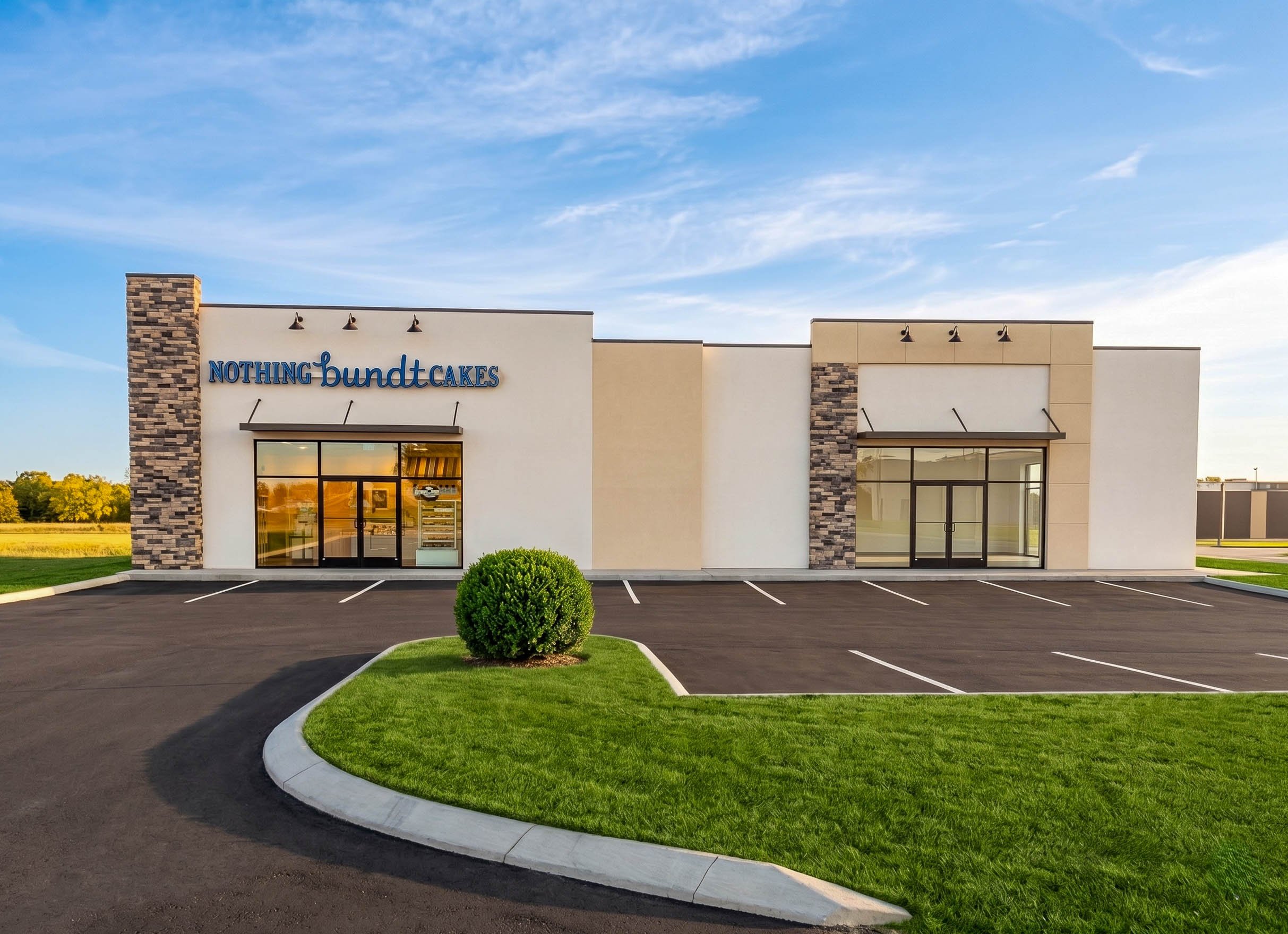 Exterior of a bakery named 'Nothing Bundt Cakes' with a parking lot, green lawn, and a clear blue sky.