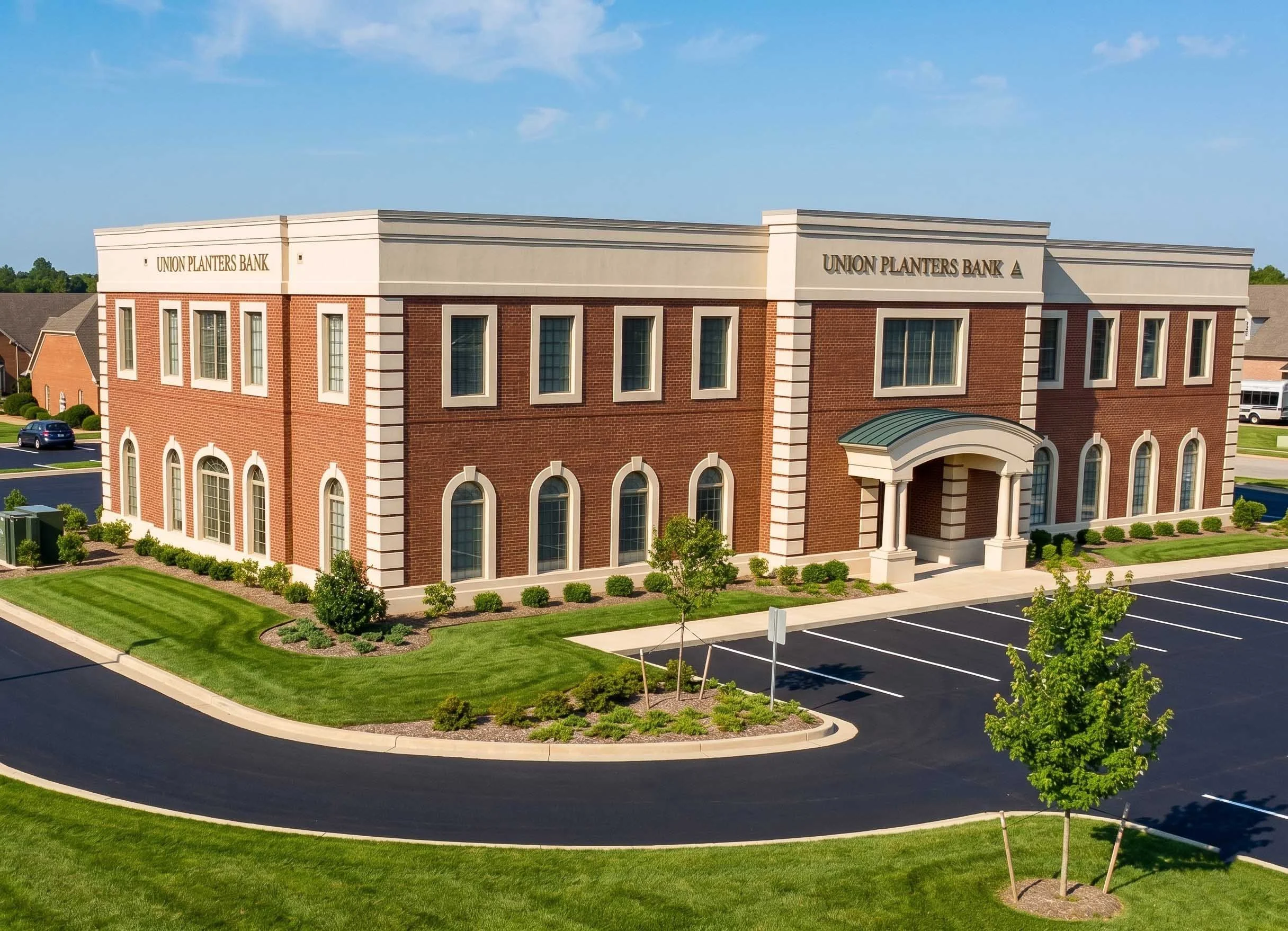 A two-story brick bank building with a white sign reading 'Union Planters Bank' on top, surrounded by a parking lot, green lawns, and trees, under a blue sky with a few clouds.