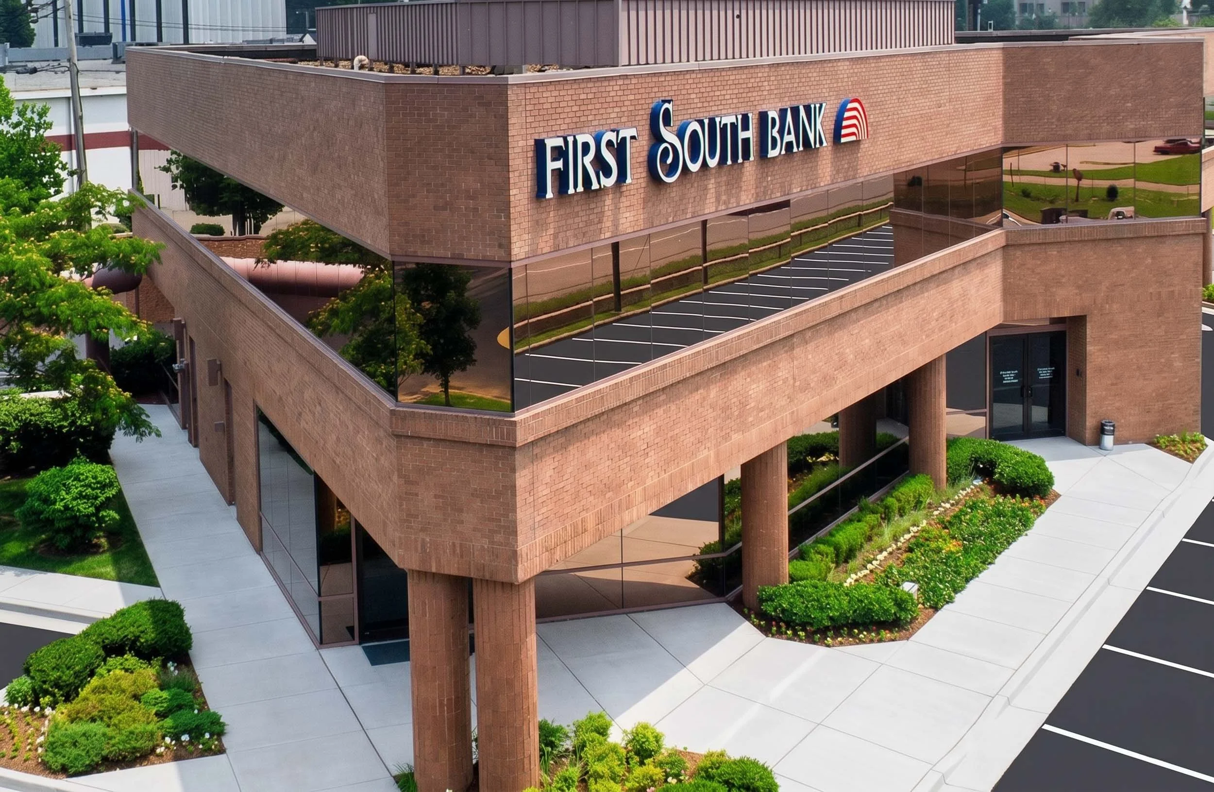 A multi-story brick building with reflective glass windows and a sign that reads 'First South Bank' on the top floor. The building is surrounded by a landscaped area with green shrubs, trees, and a sidewalk, and there is a parking lot nearby.
