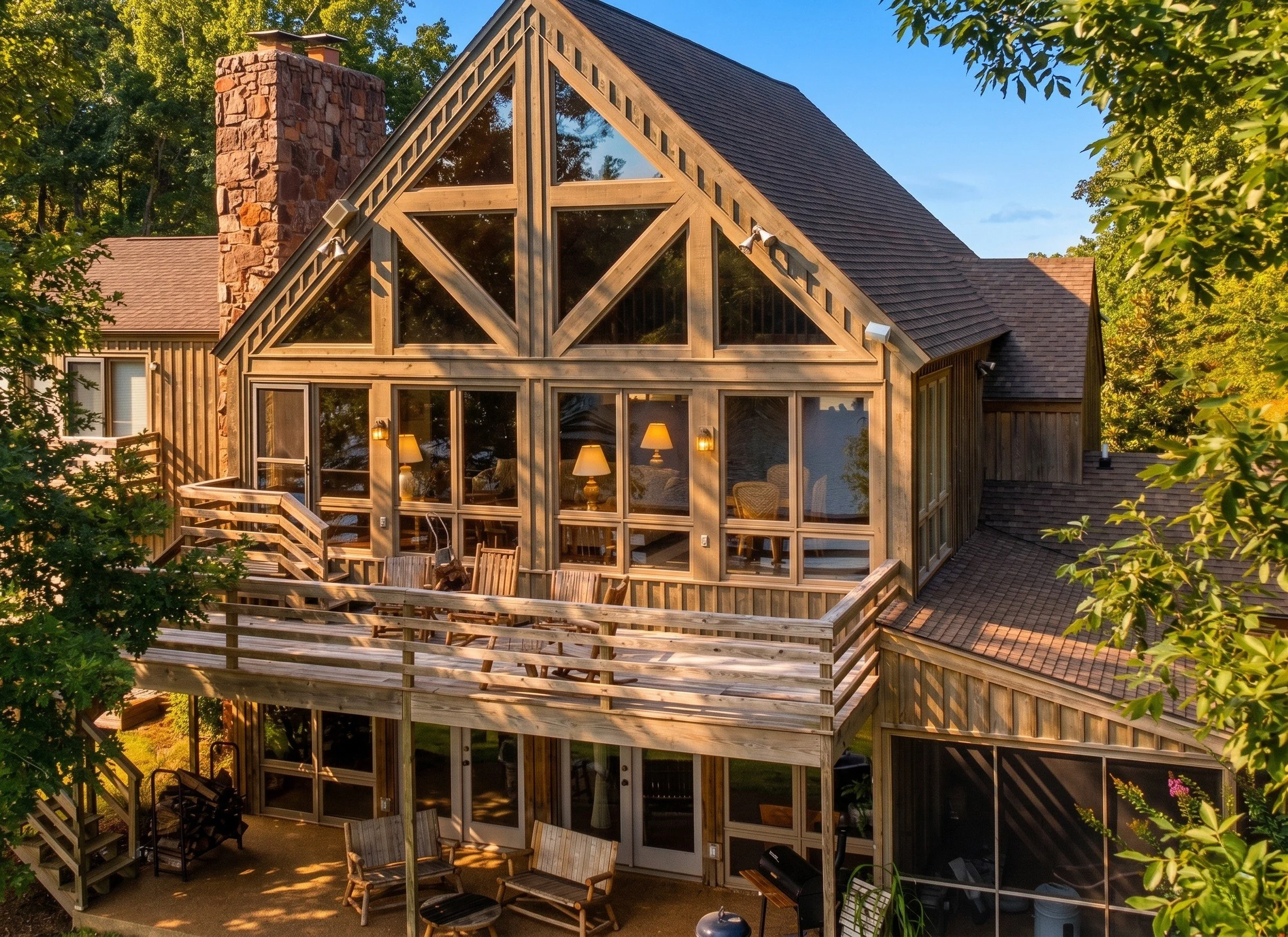 A house with a large glass windowed gable facing the backyard, featuring a wooden deck with outdoor furniture, surrounded by greenery and trees.