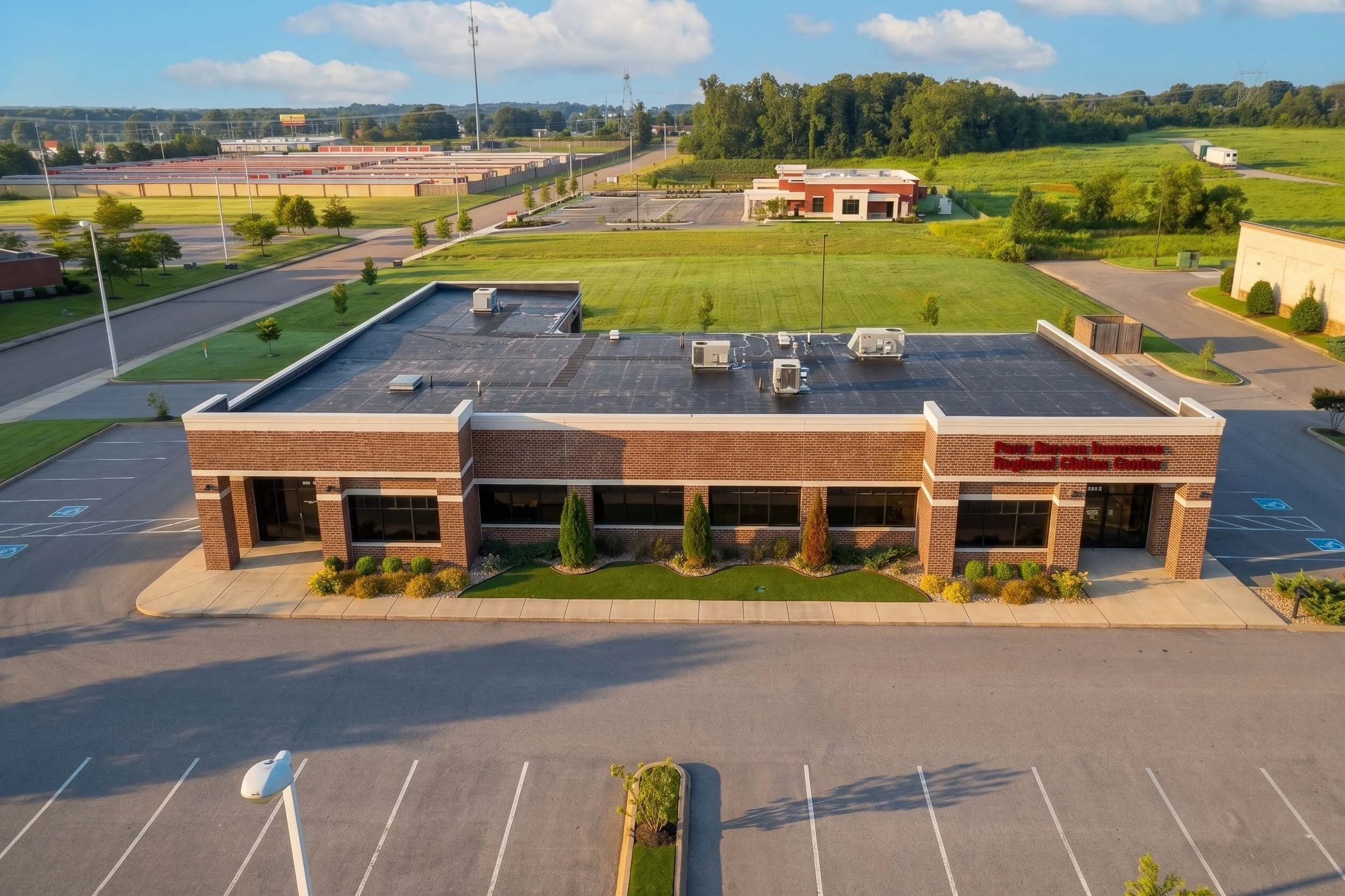 An aerial view of a commercial building with solar panels on the roof, surrounded by parking lot and green landscaping, with a grassy field, trees, and distant roads in the background.