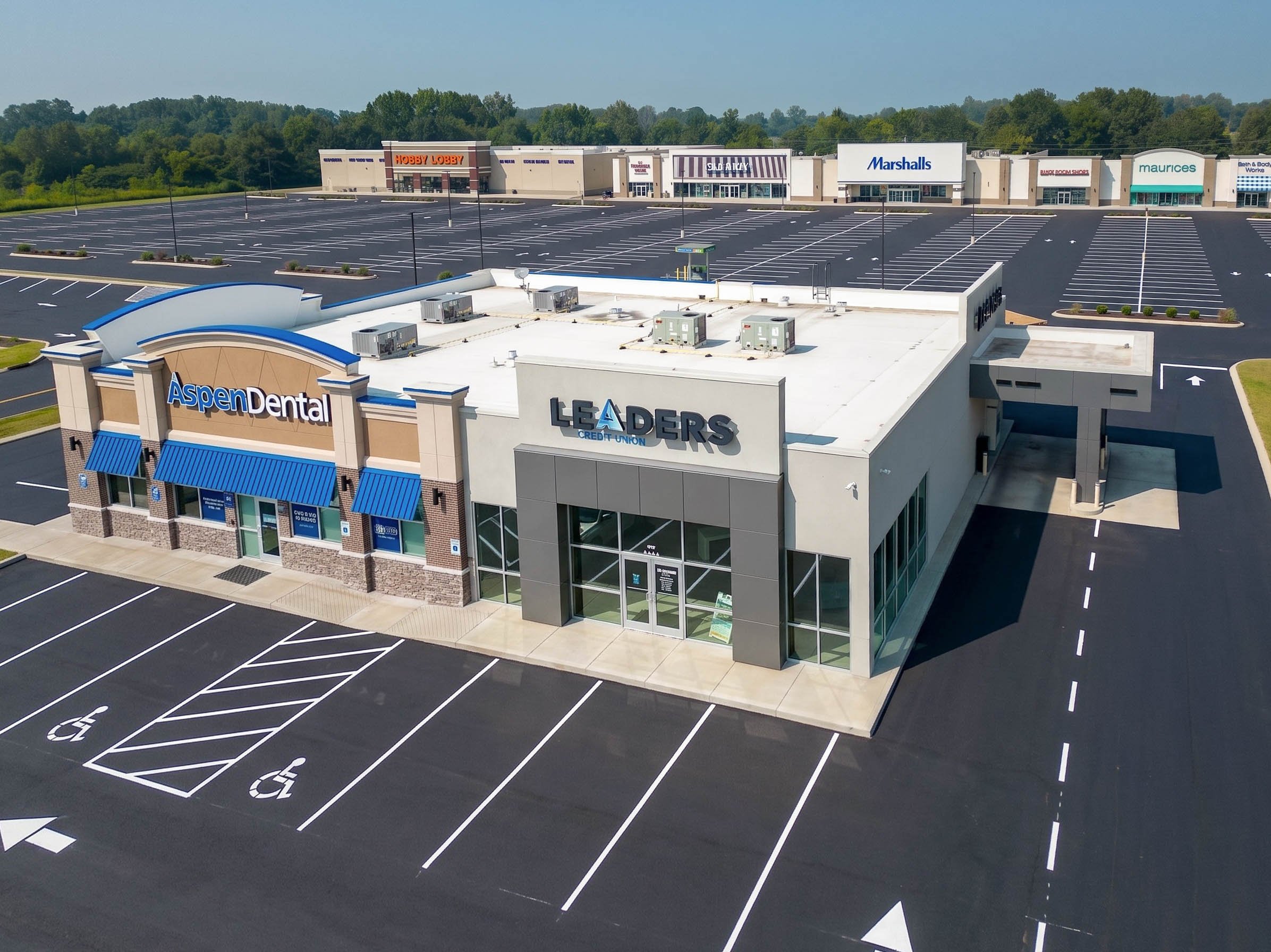 Empty parking lot outside a shopping plaza with stores including Aspen Dental, Leaders Credit Union, Marshalls, and Maurice's, under a clear sky surrounded by green trees.