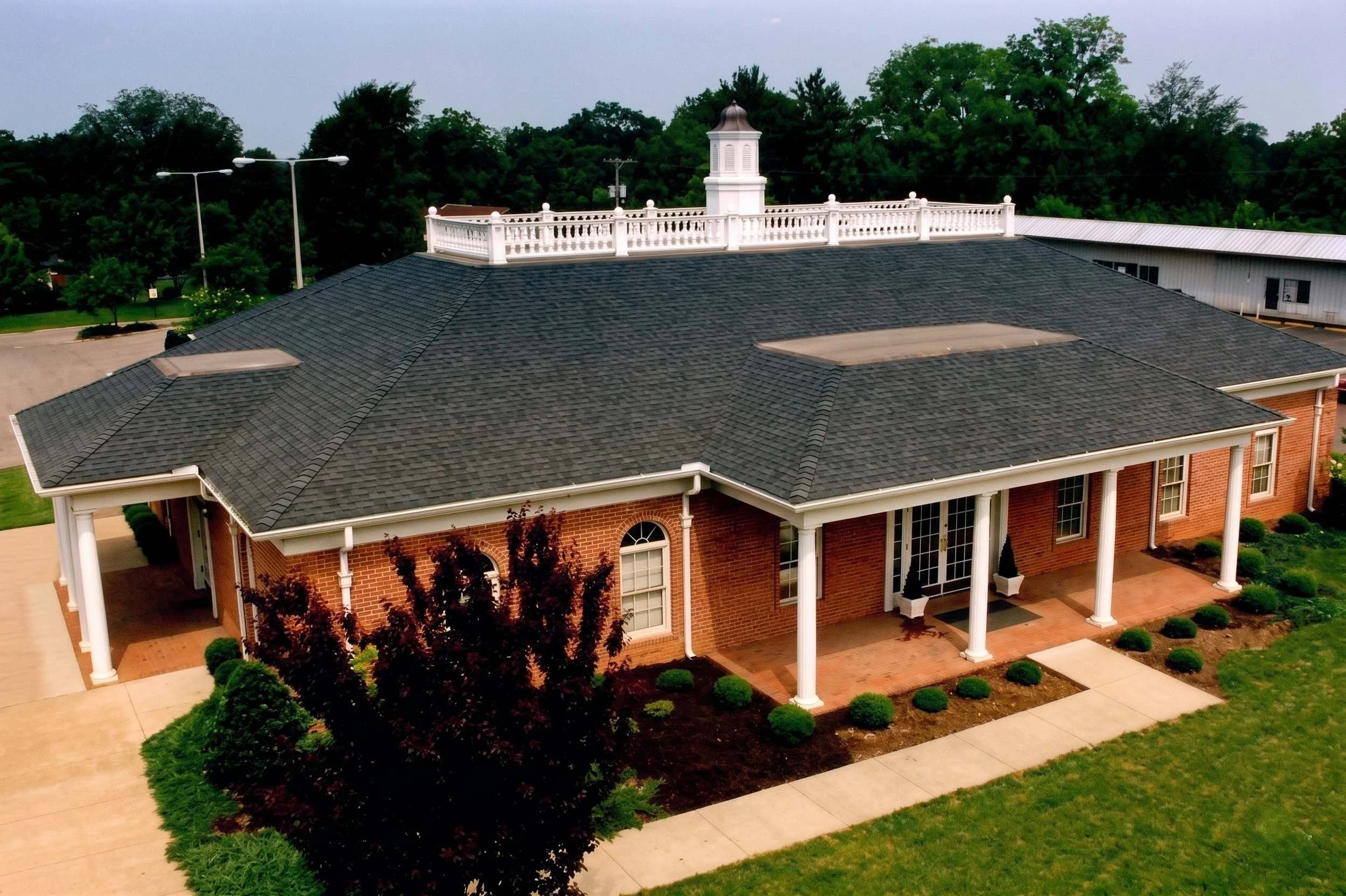 An aerial view of a brick building with a dark shingle roof, a white balustrade on top, and surrounding landscaped greenery including bushes, trees, and a concrete sidewalk.