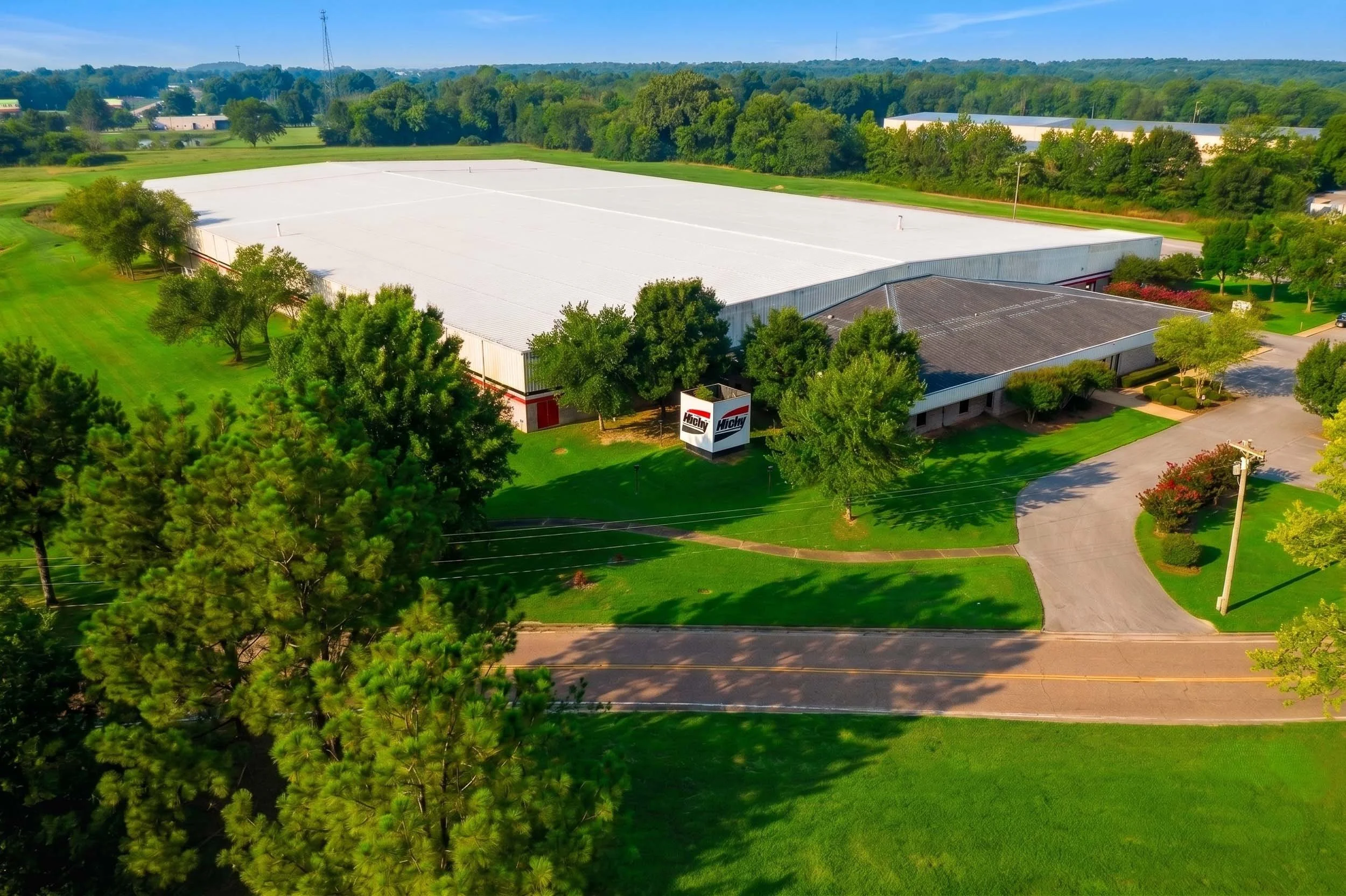 Aerial view of a large industrial or warehouse building with a white roof, surrounded by green trees and landscaped lawns, with a parking lot and road in the foreground.