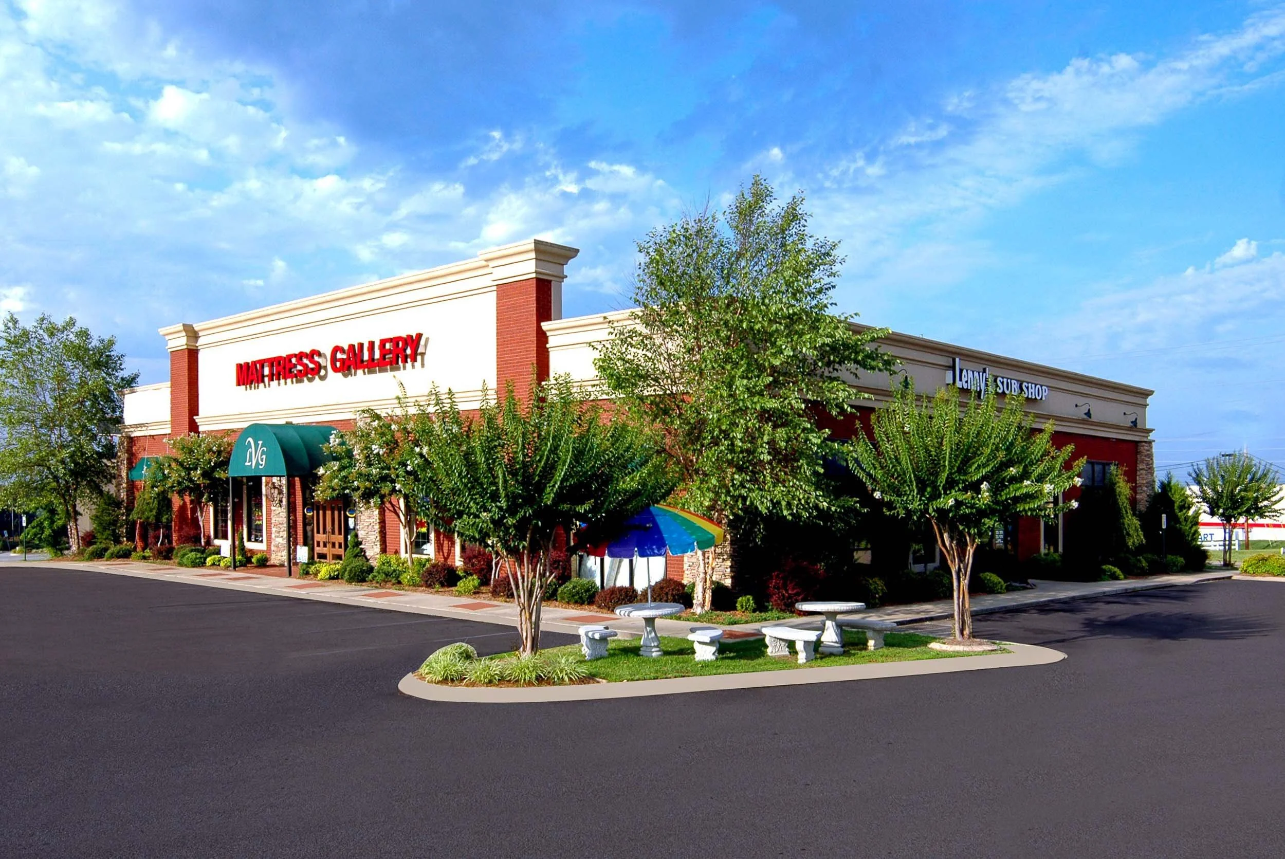 A shopping plaza with a sign for Mattress Gallery and Lenny's Sub Shop, surrounded by trees and a parking lot with a couple of picnic tables and an umbrella.
