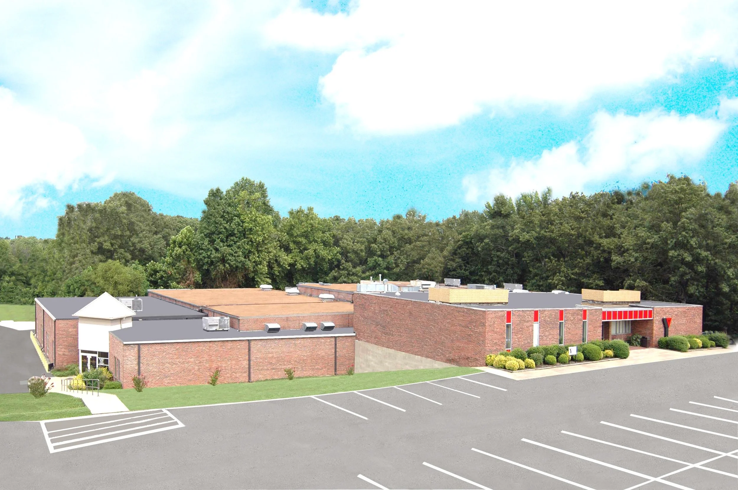 A parking lot with empty spaces in front of a red brick building, surrounded by green bushes and trees, with a blue sky and some white clouds overhead.