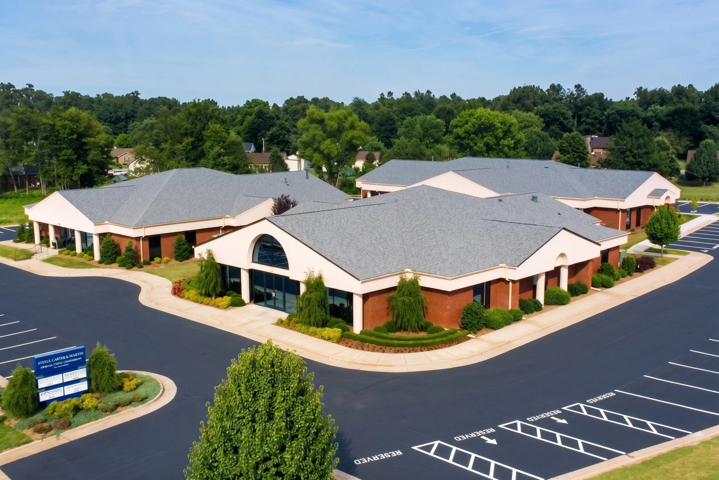 Aerial view of a commercial building with a gray roof, surrounded by parking lot spaces, green trees, and a lush wooded background.