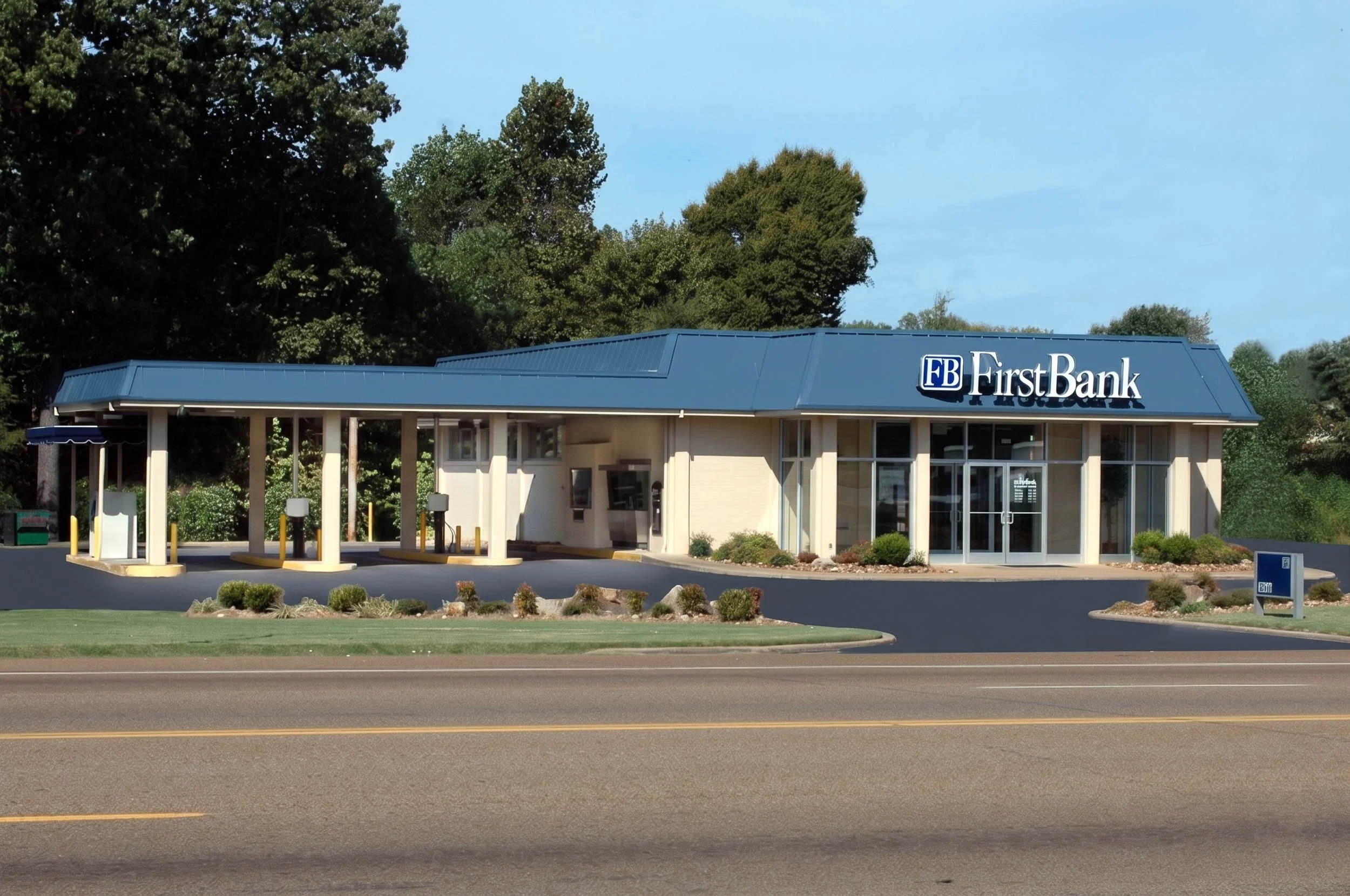 A small building with a blue roof and a sign that says FirstBank. There are parking lots in front, surrounded by some landscaped bushes and trees in the background, under a clear blue sky.