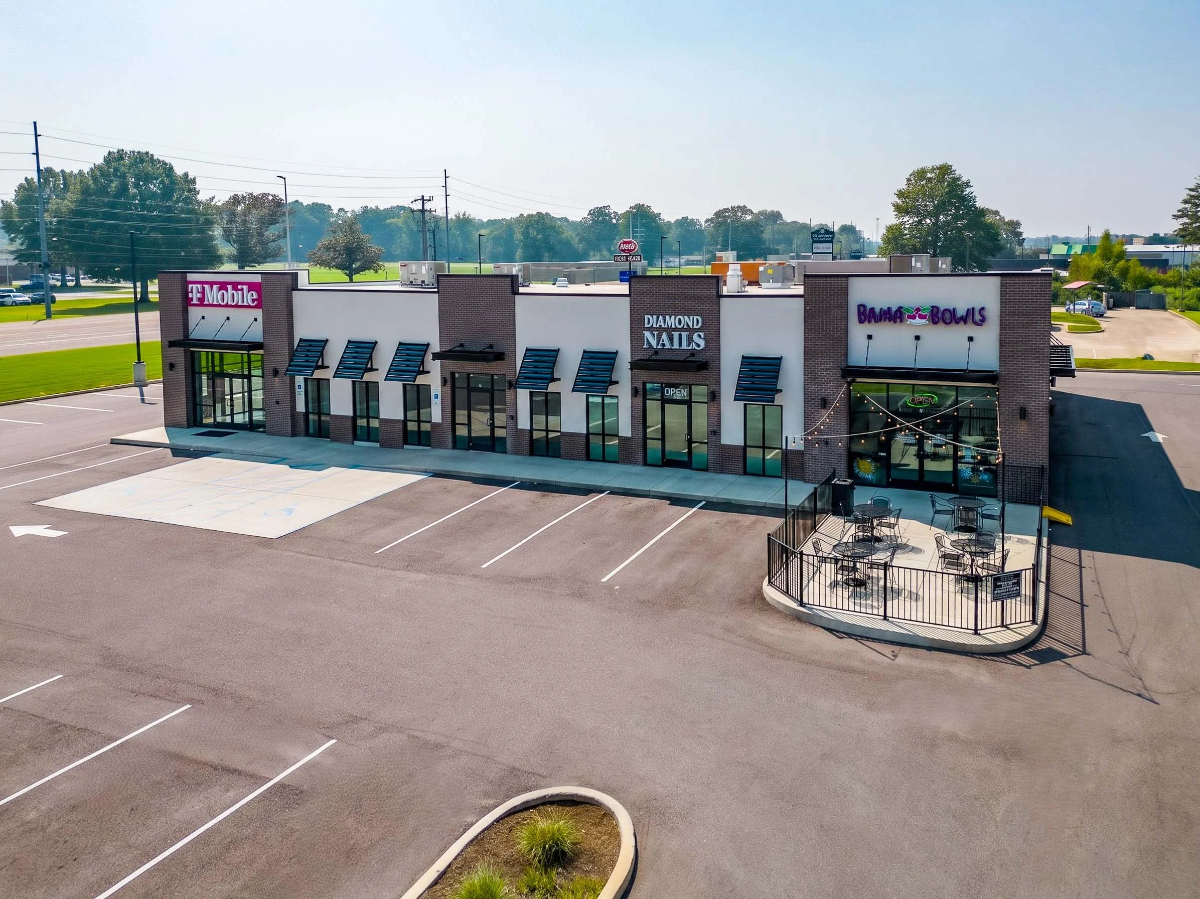 A shopping plaza with three storefronts: T-Mobile, Diamond Nails, and Bahama Bowls. The parking lot is empty with clearly marked parking spaces, and there are small planters with greenery.