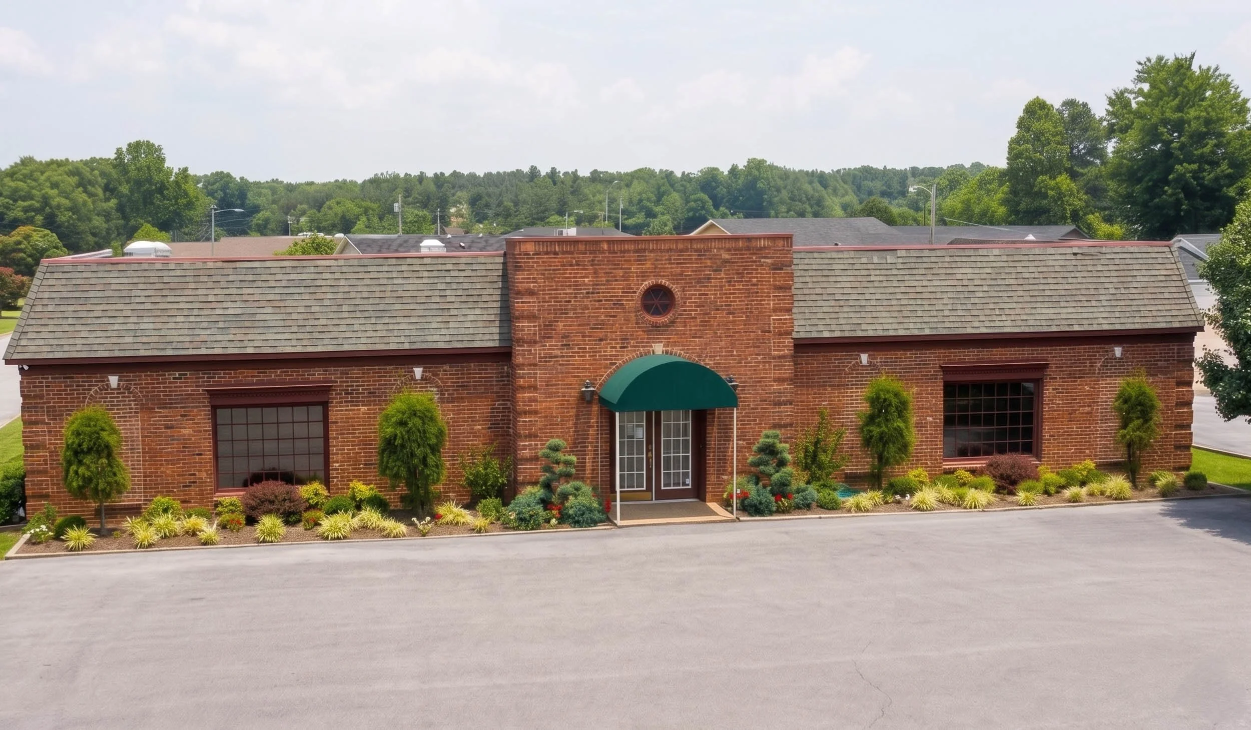 Front view of a brick building with large windows, a green awning over the entrance, and well-maintained landscaping including small trees and bushes. In the background, there are trees, rooftops, and a partly cloudy sky.
