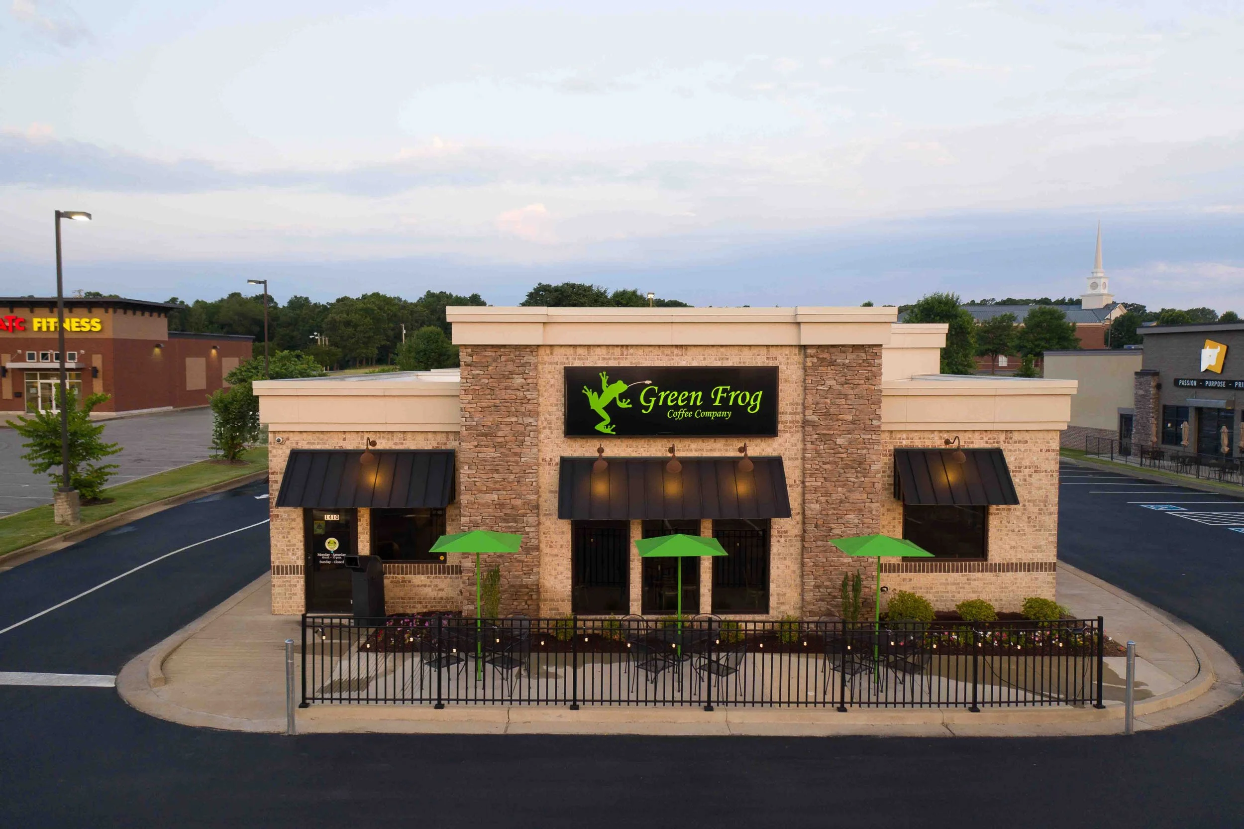 A small commercial building with a sign reading Green Frog Coffee Company, featuring a green frog logo, black awnings, outdoor seating with green umbrellas, and a parking lot in front.