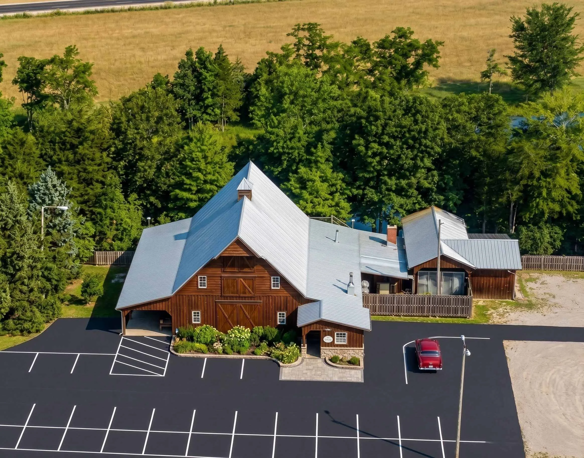 An aerial view of a rustic wooden building with a metal roof, surrounded by trees and a parking lot with a few cars, set against a background of grassy fields.