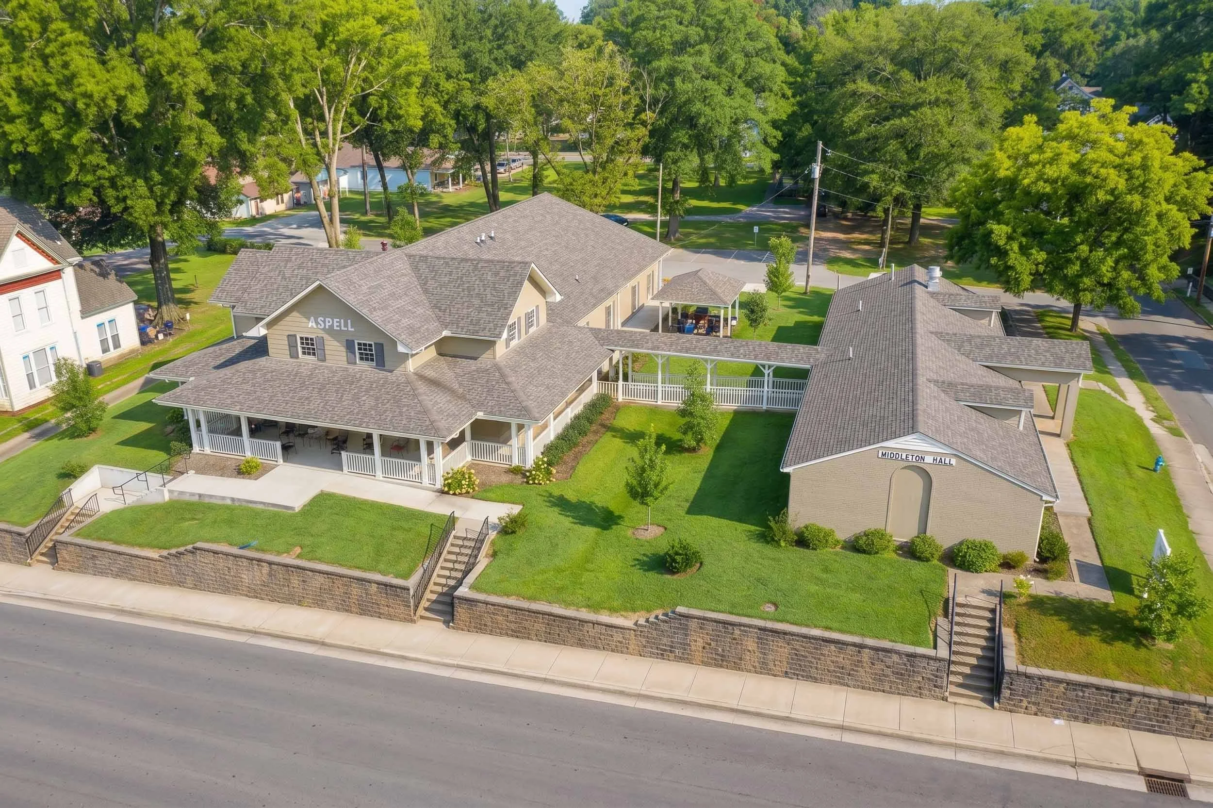 An aerial view of a residential neighborhood showing a house with a porch, trees, and a surrounding yard, with a street in front and a stone wall along the sidewalk.