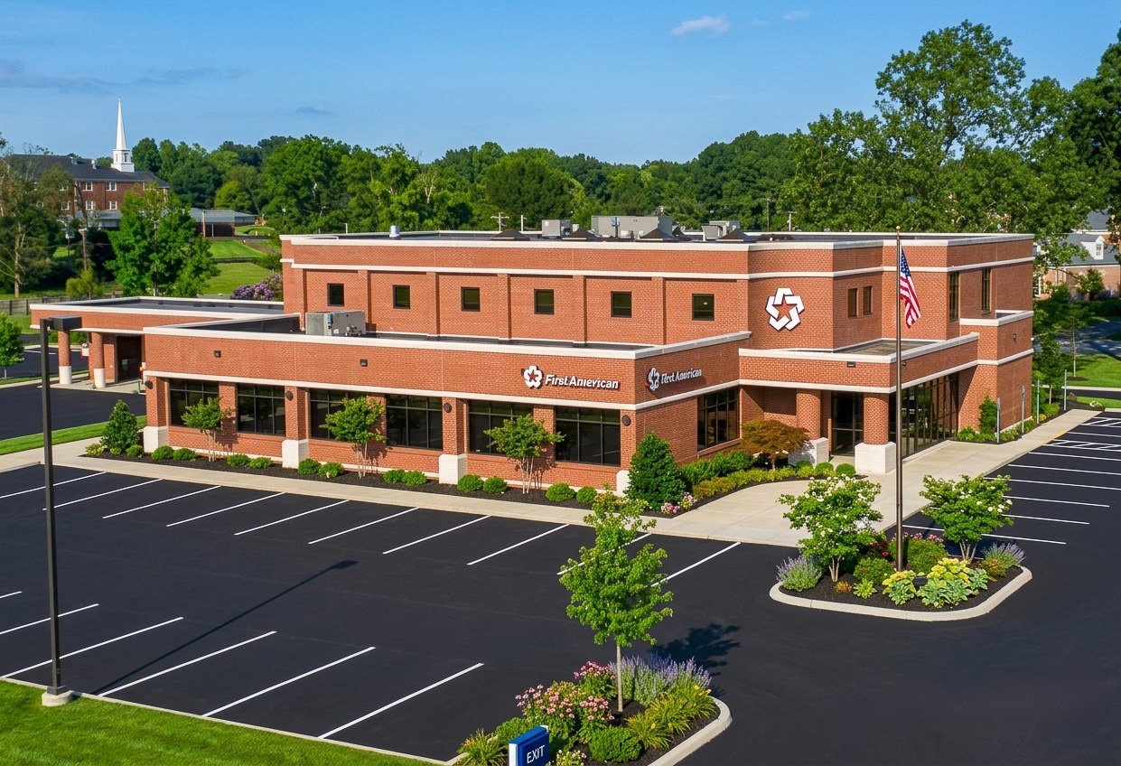 A modern red brick building with large windows and is surrounded by a landscaped parking lot with trees, shrubs, and flowers. An American flag is flying on a pole near the entrance, and a white steepled church is visible in the background amidst gree