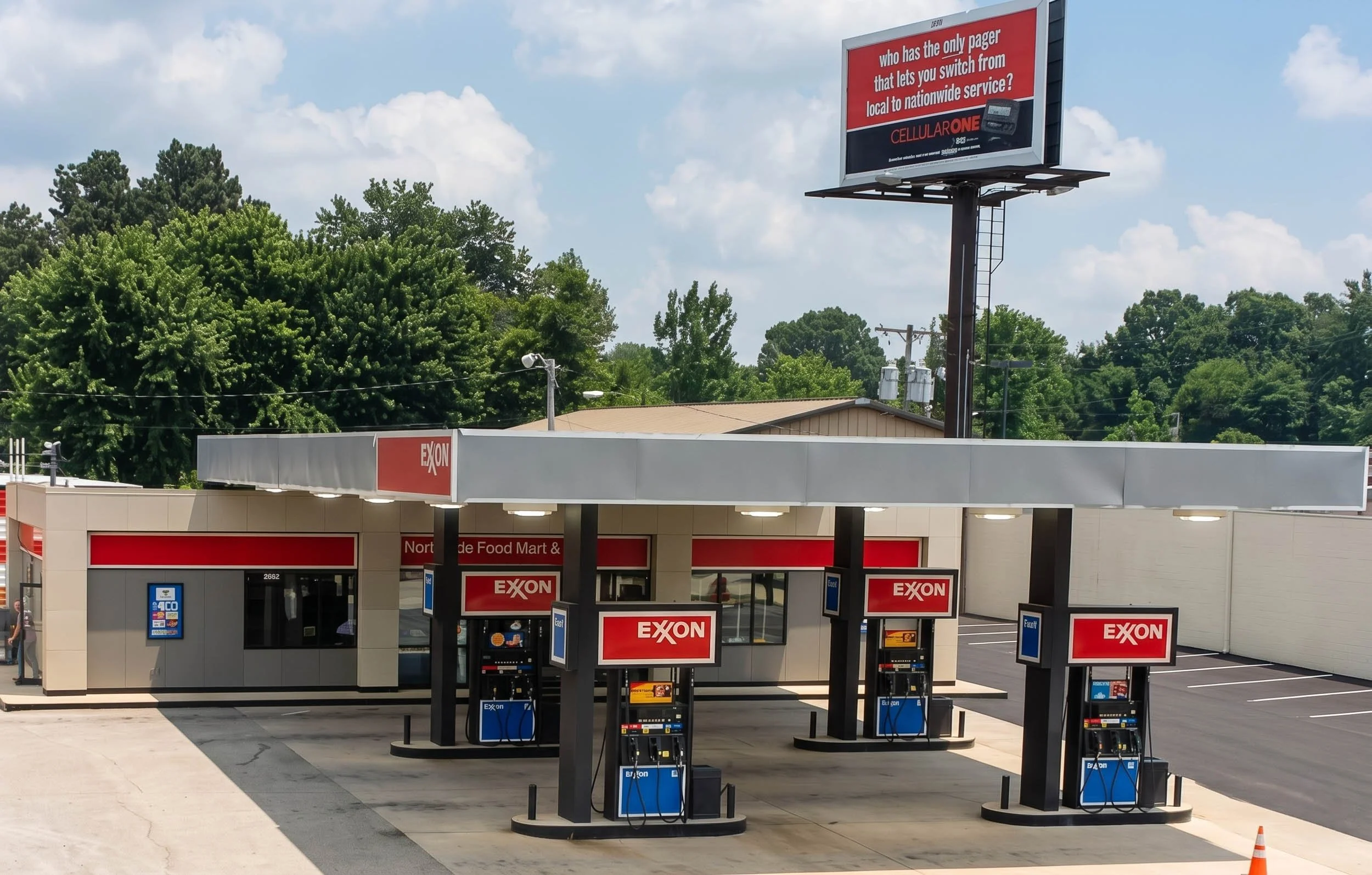 A gas station with multiple gasoline pumps branded with Exxon logos, and a large billboard advertising cellular phone service. The station is outdoors with trees and a partly cloudy sky in the background.