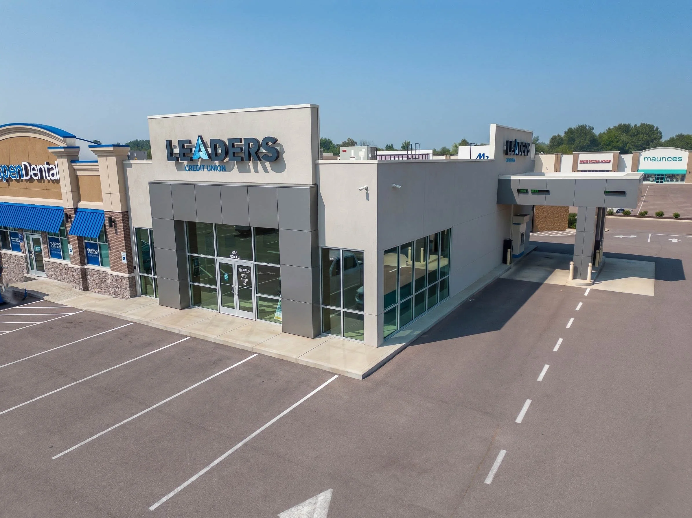 Empty parking lot with marked parking spaces in front of a shopping center on a clear day, featuring stores like Leaders Credit Union, Open Dental, and Maurices.