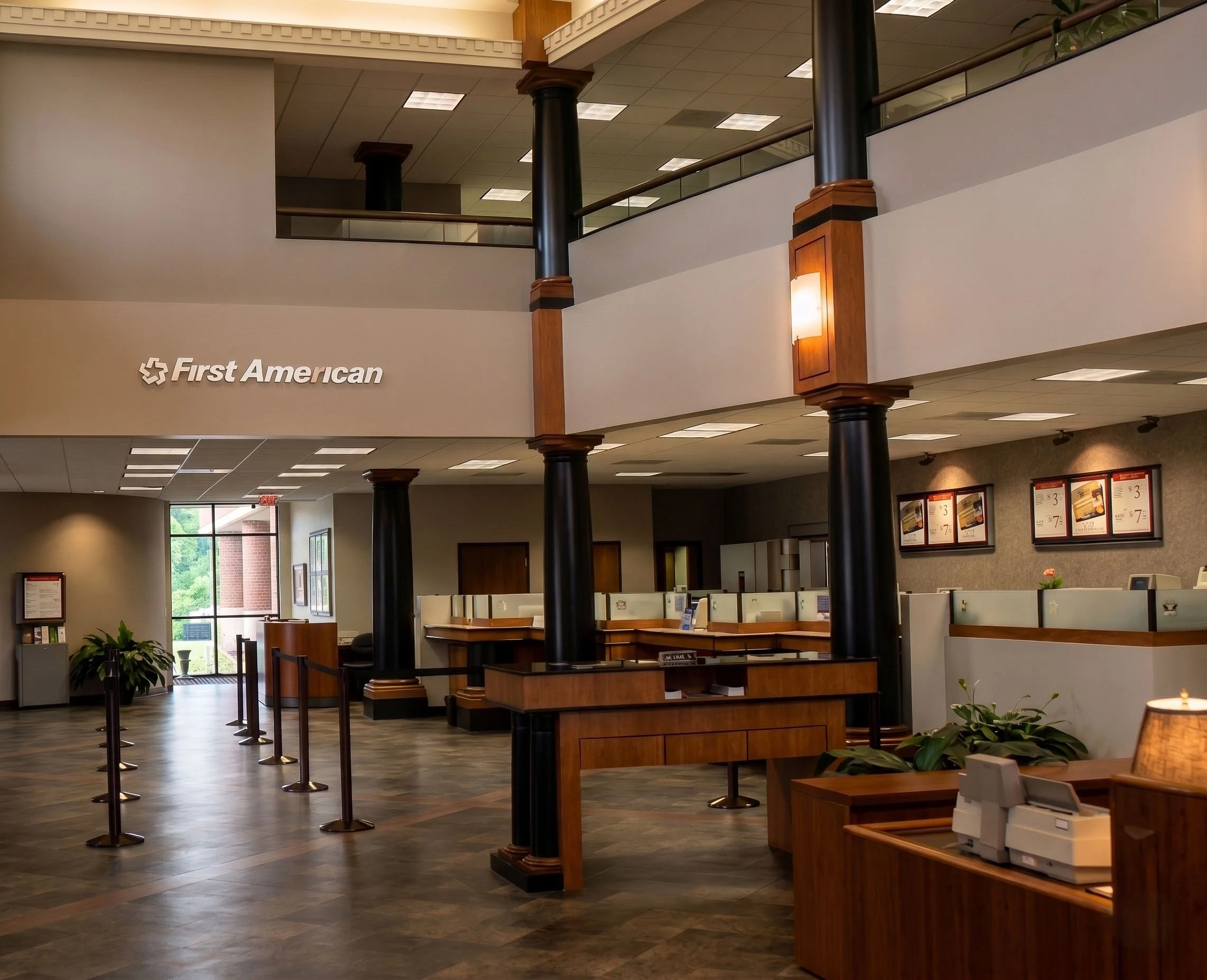 Interior of a First American bank branch with teller stations, a waiting area, plants, and informational display screens.