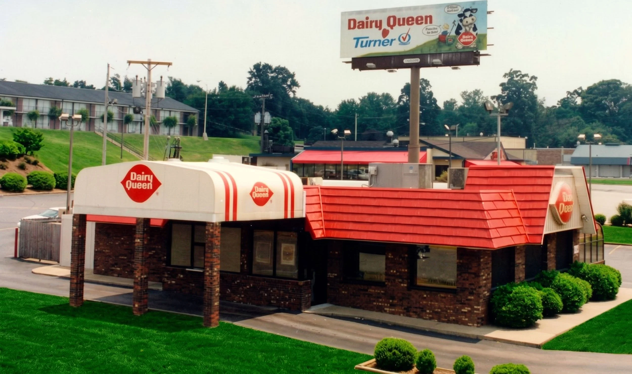 Dairy Queen fast food restaurant with red roof and brick exterior, surrounded by manicured green lawn and bushes, with a large billboard advertising Dairy Queen in the background.