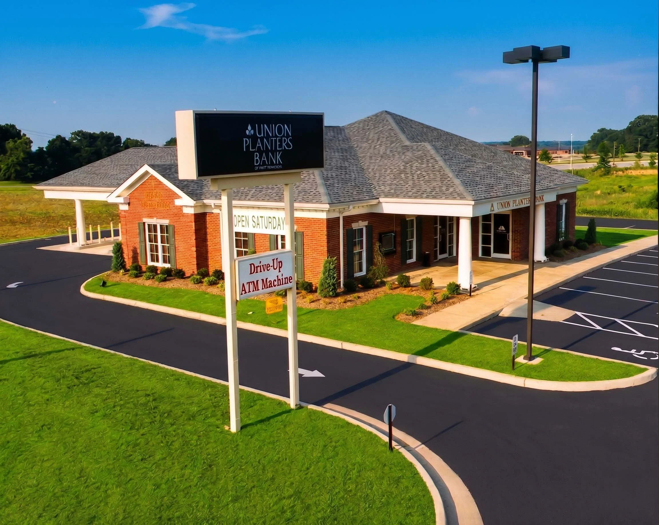 A brick building with a sign that reads 'Union Planters Bank.' There is a parking lot with spaces marked and a tall light pole nearby. A green lawn surrounds the building, and there is a smaller sign advertising drive-up ATM services. The sky is clea
