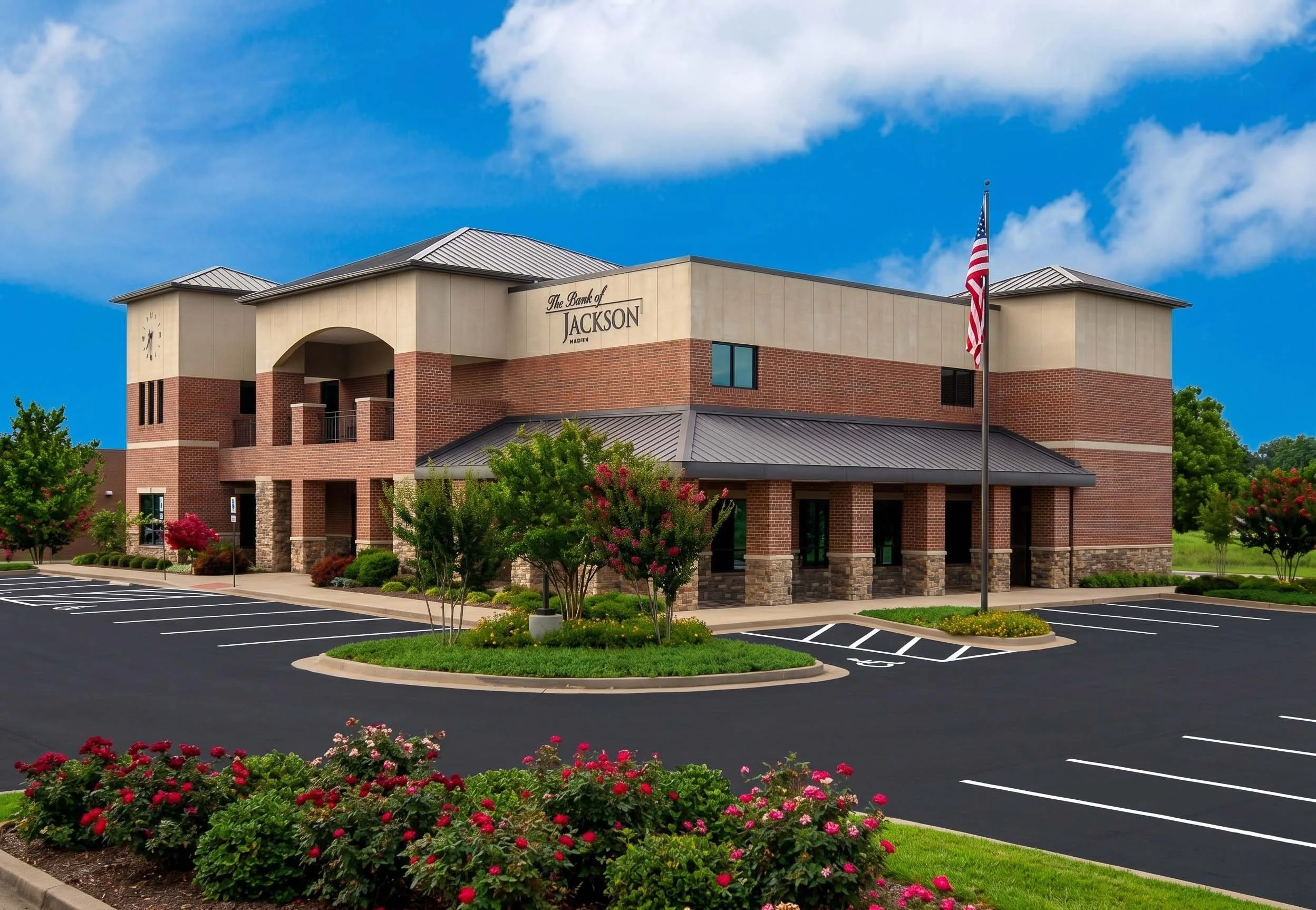 A modern brick and beige building labeled 'The Bank of Jackson' with a parking lot and landscaped trees and flowers in the foreground, with a blue sky and clouds in the background.