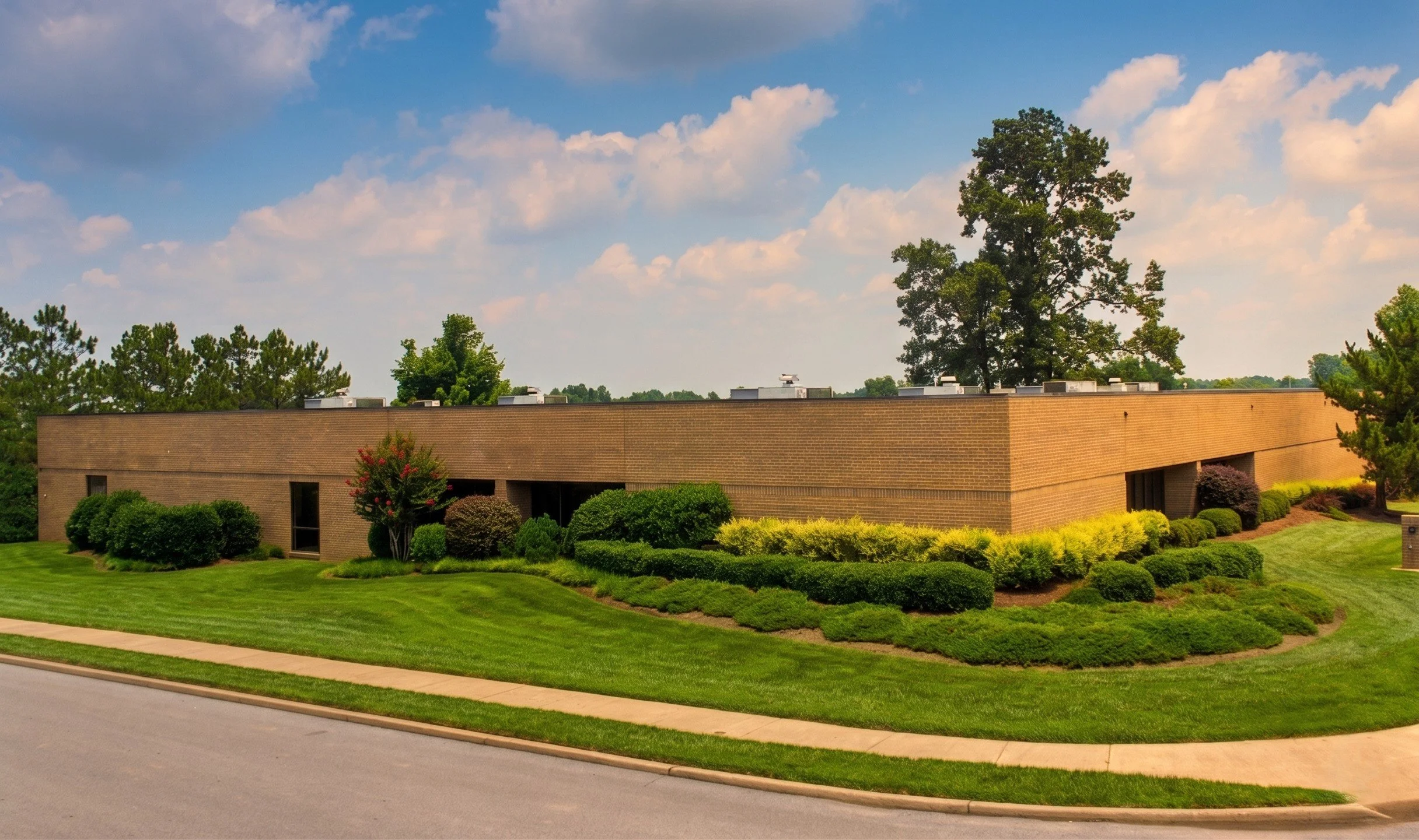 A beige brick building with a flat roof surrounded by green bushes, trees, and a well-maintained lawn under a partly cloudy sky.