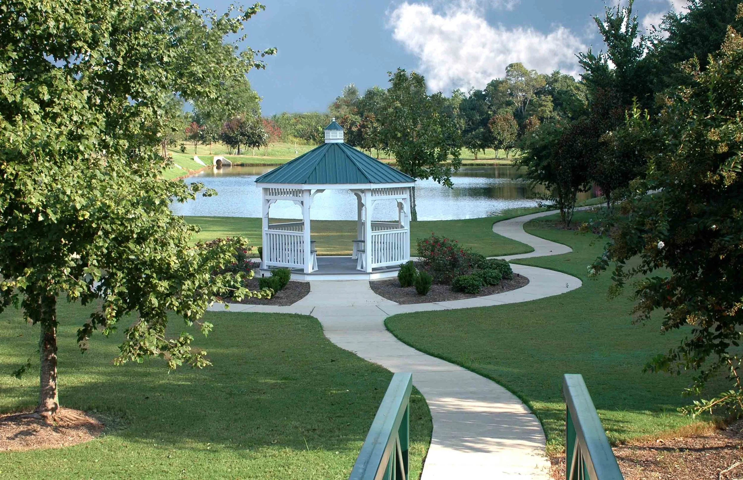 A park scene with a white gazebo with a blue roof, a winding pathway, lush green grass, trees, a pond, and a bridge.