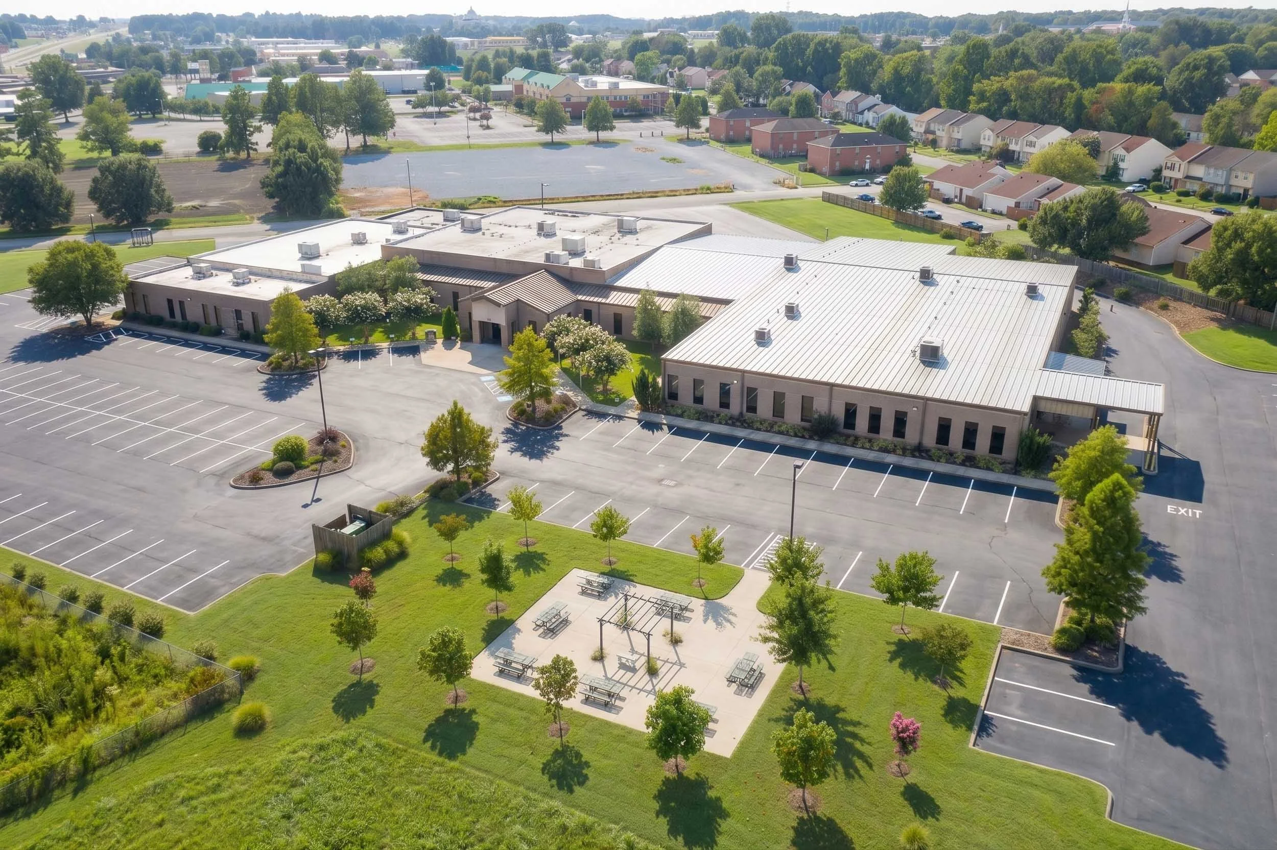 An aerial view of a school campus with a large parking lot, green lawns, trees, a playground, and surrounding residential houses in a suburban area.