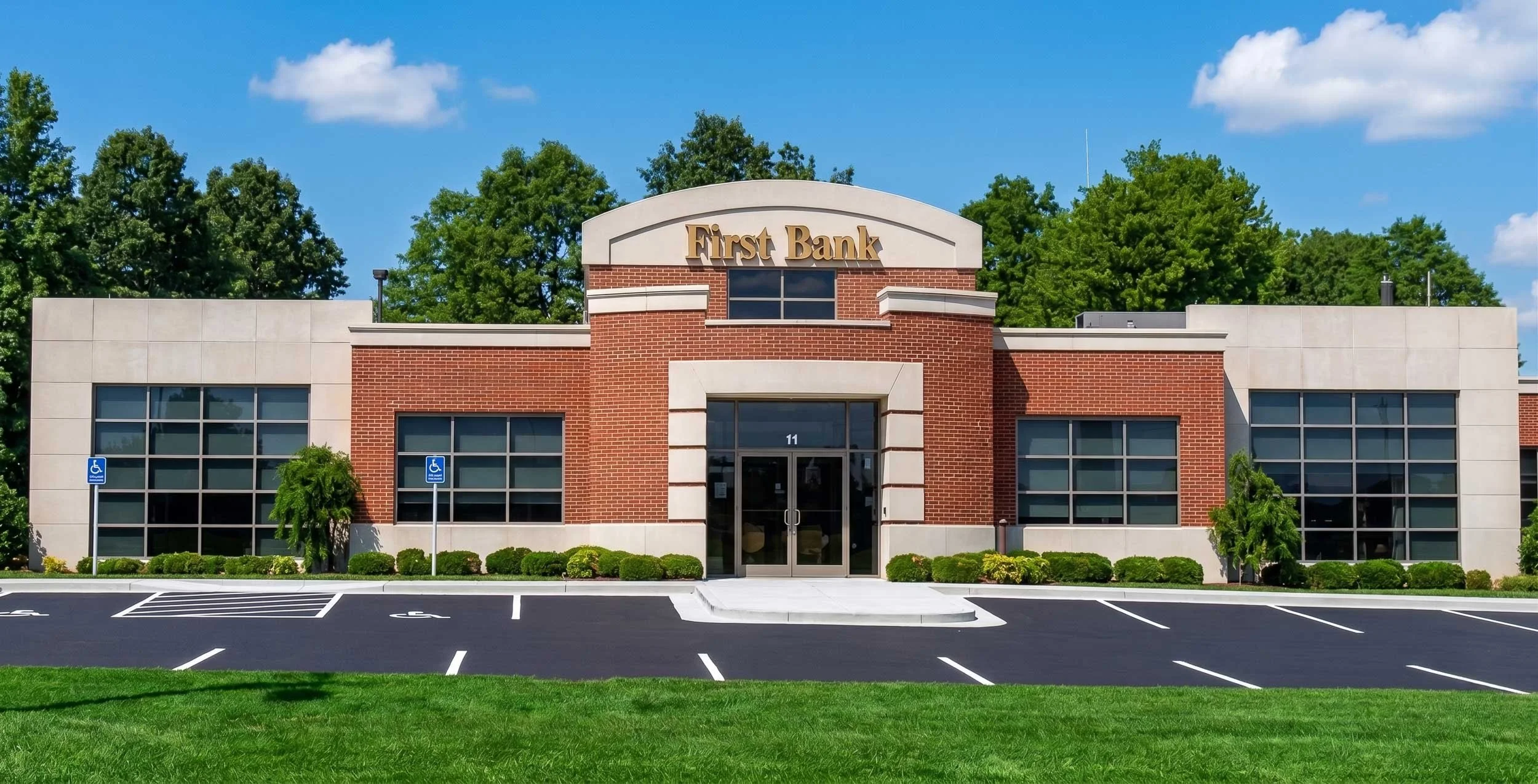The image shows a modern brick and concrete bank building with a sign that reads 'First Bank' above the entrance. The bank has large windows, and there are parking spaces in front, including designated handicap spots. There is well-maintained grass a