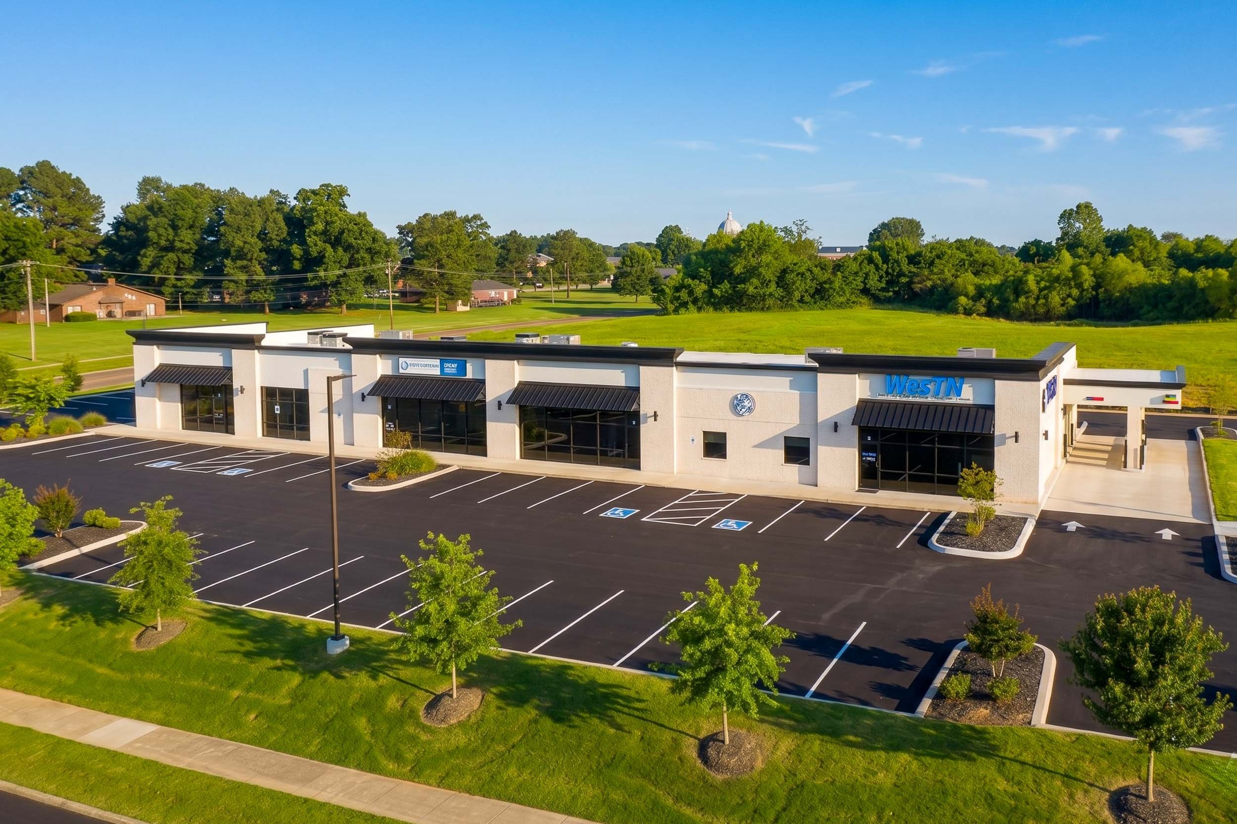 A single-story commercial building with a parking lot and trees, situated in a green landscape under a clear blue sky. The building has a sign that reads "WestN" and features black awnings over large windows.