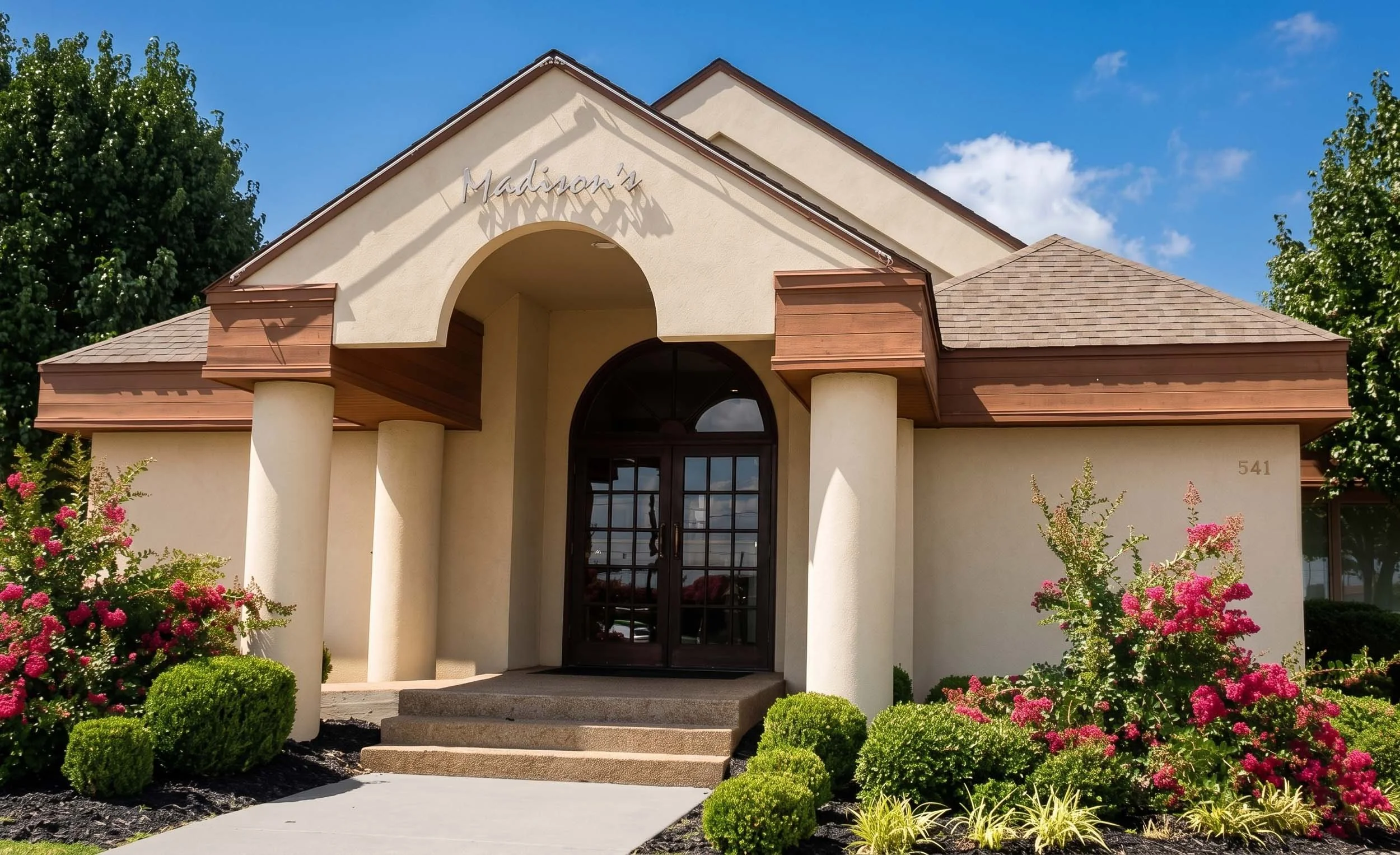 Front view of a light-colored building with a gabled roof, two large white pillars, and a curved entrance with glass doors. The building has a sign that reads 'Madison's' above the entrance, and the address 541 is visible on the wall. The landscape i