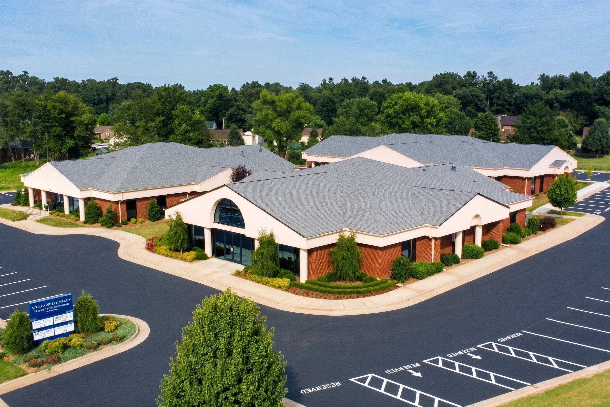 An aerial view of a commercial building with a grey roof, surrounded by a parking lot and landscaped plants, with trees and a blue sky in the background.
