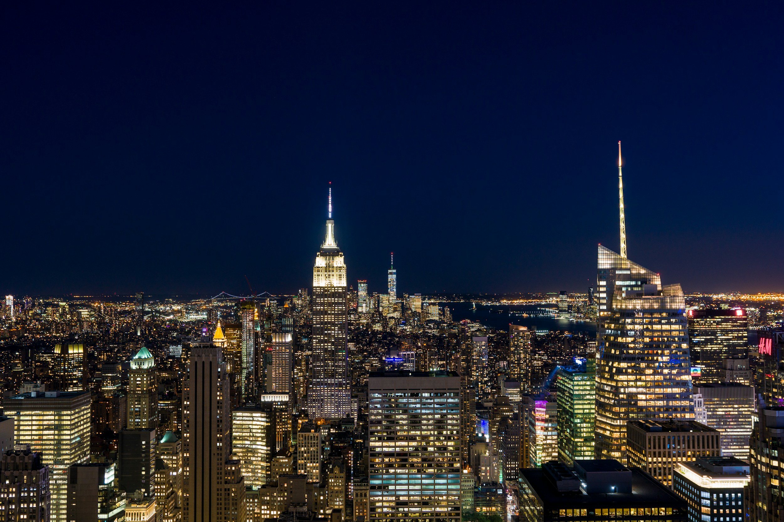 Night view of the New York City skyline featuring the Empire State Building and other illuminated skyscrapers, with a dark sky overhead.
