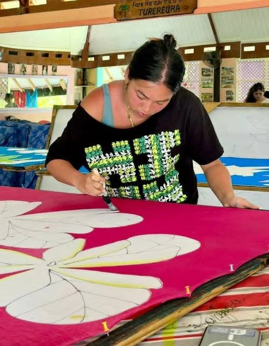 A woman painting a large flower on a red fabric at a workshop or art event in a tropical setting.
