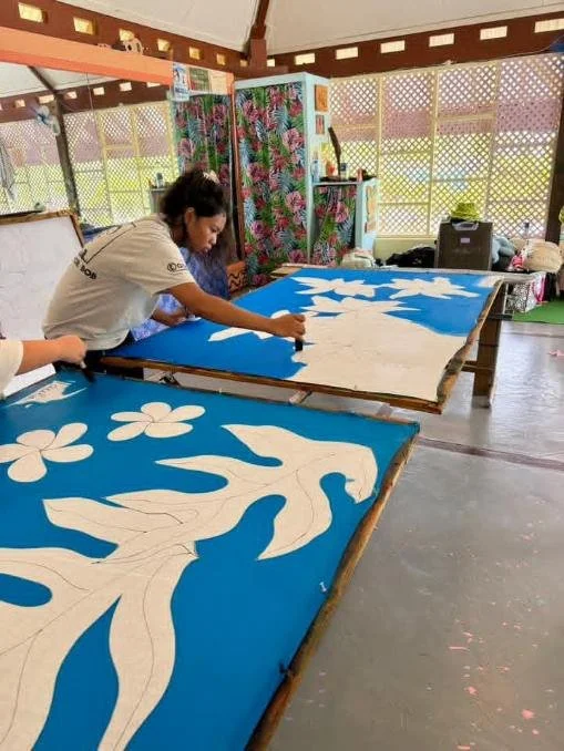 Person working on large blue and white fabric designs with floral patterns in a bright room with windows and colorful curtains.