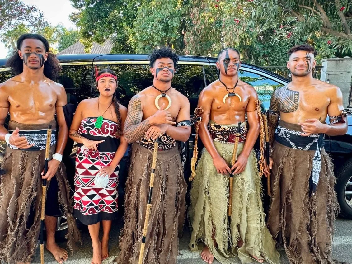 Group of five people dressed in traditional Polynesian tribal attire, including skirts and face paint, standing outdoors in front of a black vehicle, with trees in the background.