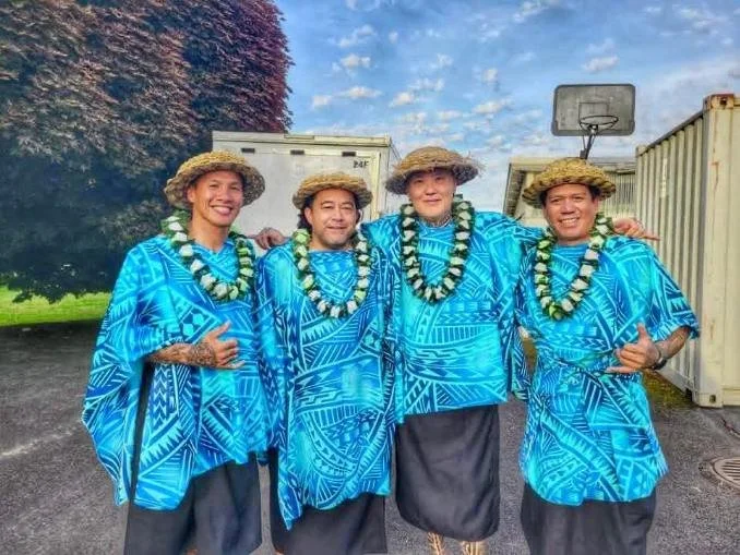 Four people in traditional blue Polynesian attire, wearing straw hats and leis, standing outdoors with arms around each other, smiling.