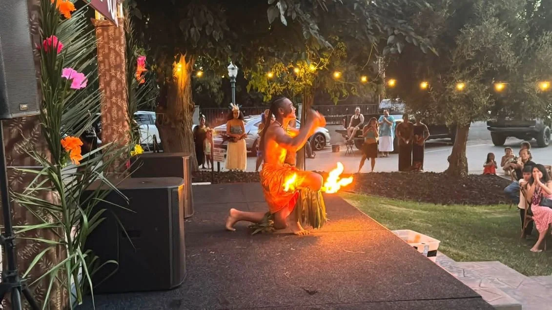 A fire performer dressed in traditional Hawaiian attire performs on an outdoor stage in front of a crowd, with trees and string lights in the background during evening time.