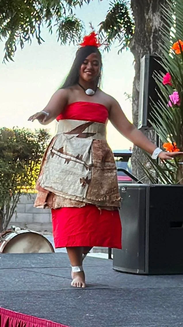 A woman dancing on stage outdoors, wearing a red strapless top, a patterned skirt, a flower headpiece, and jewelry, with tropical plants and flowers in the background.