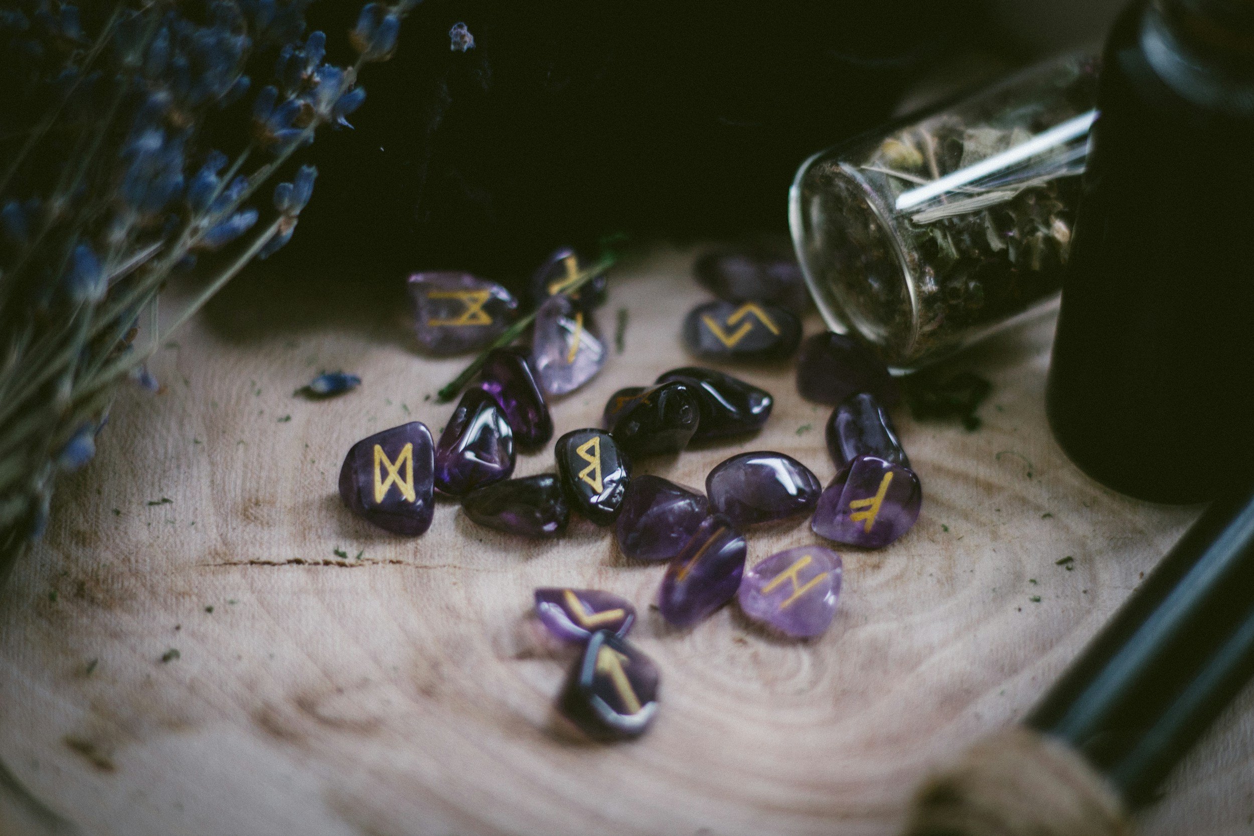 A collection of dark purple and black polished stones with yellow runes painted on them, spilled on a beige surface with a small glass jar tipped over nearby. A bunch of dried lavender is partially visible on the left side.
