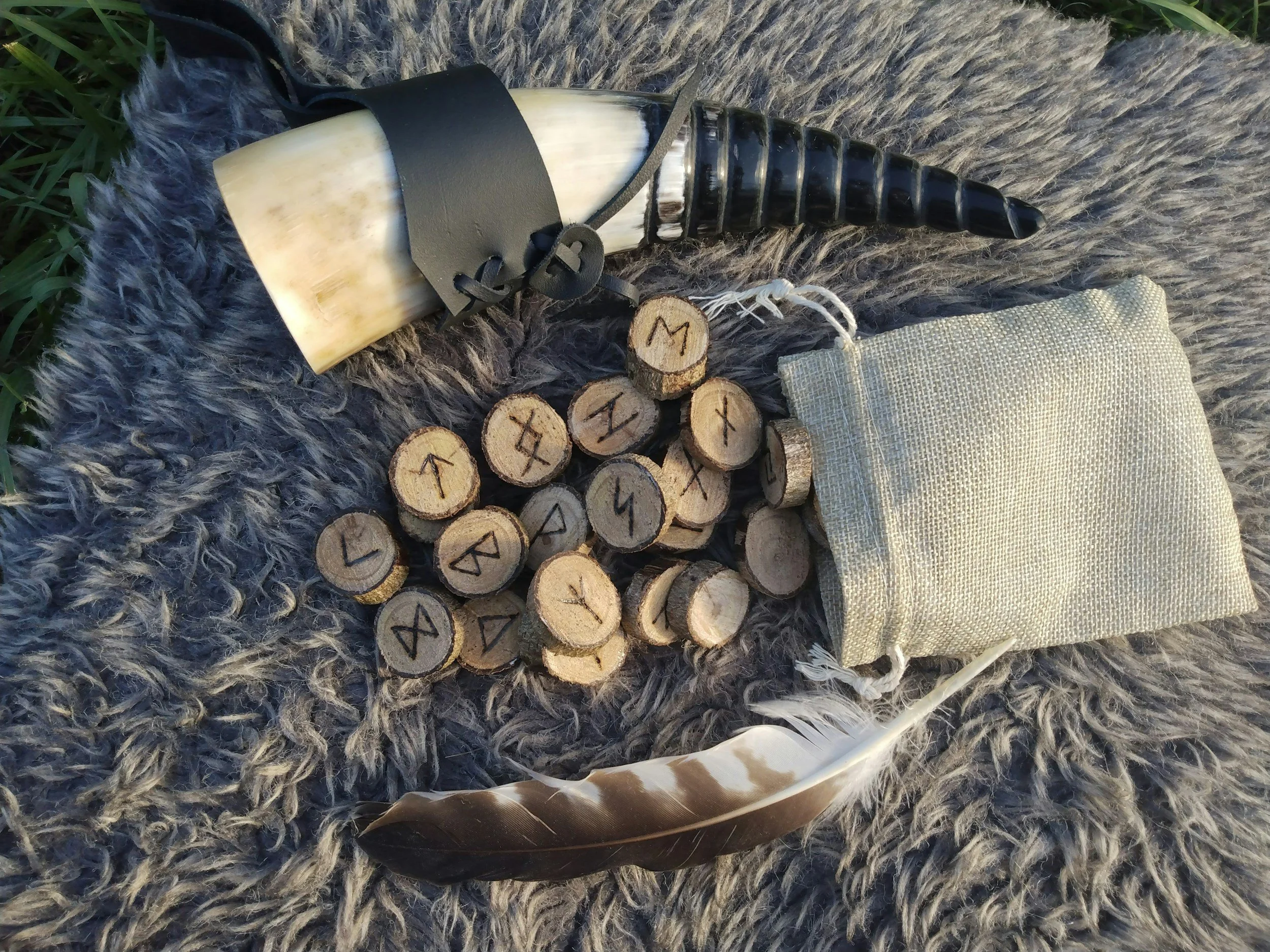 A collection of small wooden rune stones with engraved symbols, lying on a gray furry blanket. An animal horn, a beige pouch, and some feathers are also nearby.