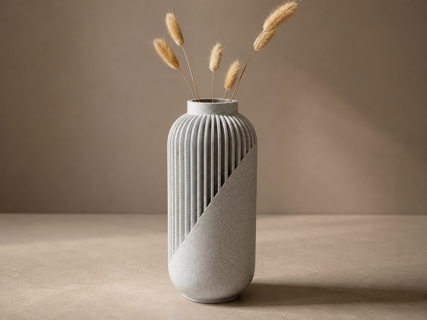 Een moderne, tweekleurige 3D geprinte vaas met verticale ribbels en een diagonale kleurverdeling, gevuld met gedroogde beige grassprieten, geplaatst op een neutraal beige oppervlak tegen een effen achtergrond.