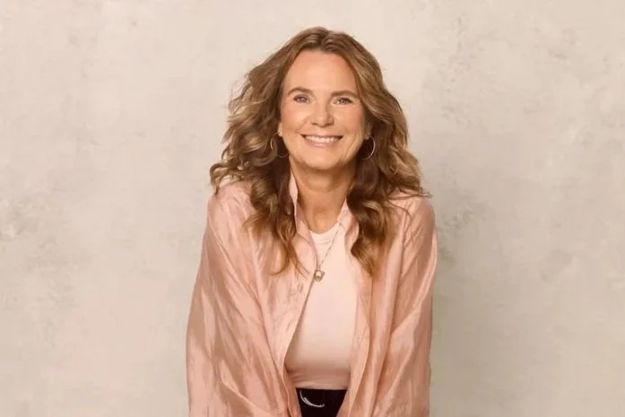 Ein lächelnder Frau mit lockigem, hellbraunem Haar, trägt ein hellrosa Jacke und ein weißes Oberteil, steht vor einem neutralen Hintergrund.