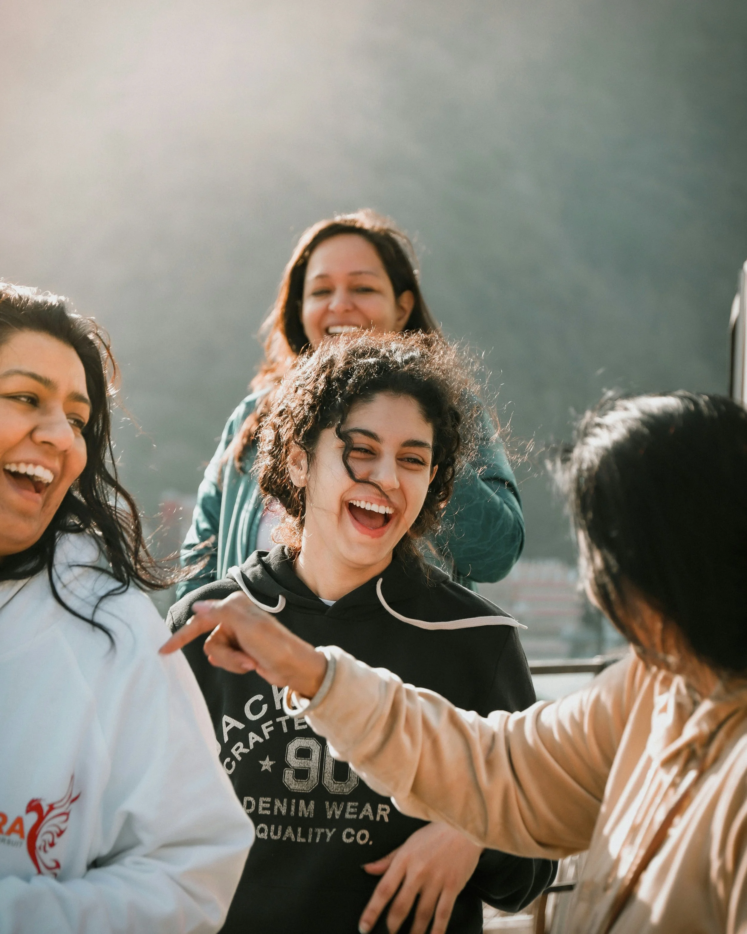 Group of smiling women hugging and laughing together outdoors with a cloudy sky in the background.