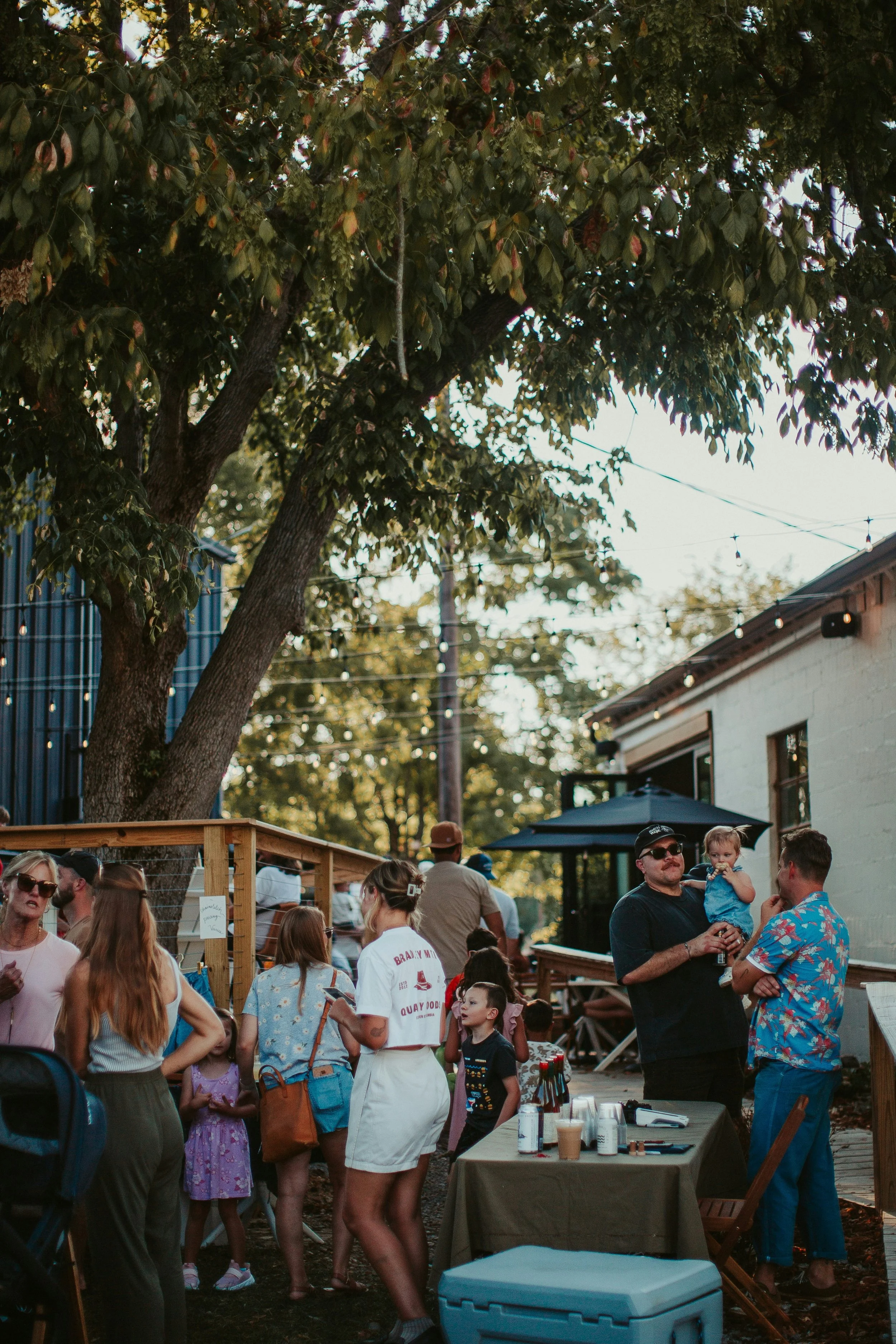 People gathered outdoors at a social event, with some standing by a table with drinks and others mingling. There are trees, string lights, and a building in the background, creating a festive atmosphere.