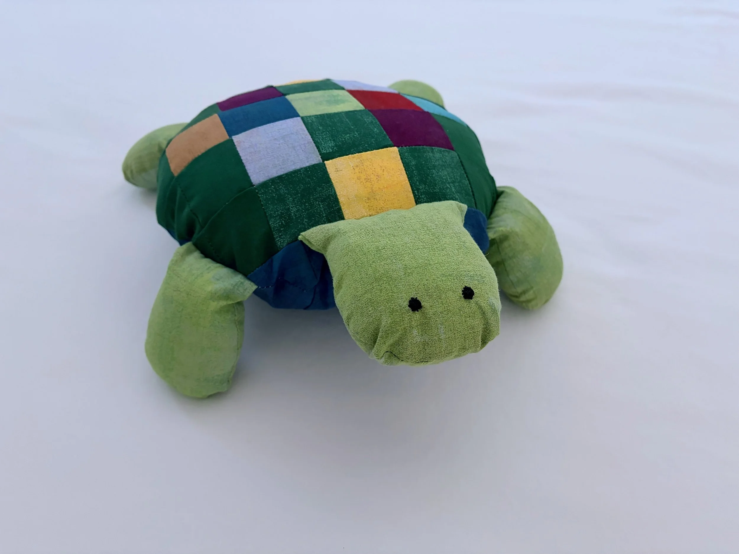 A plush turtle toy with a patchwork multicolored shell and green body, lying on a plain white surface.