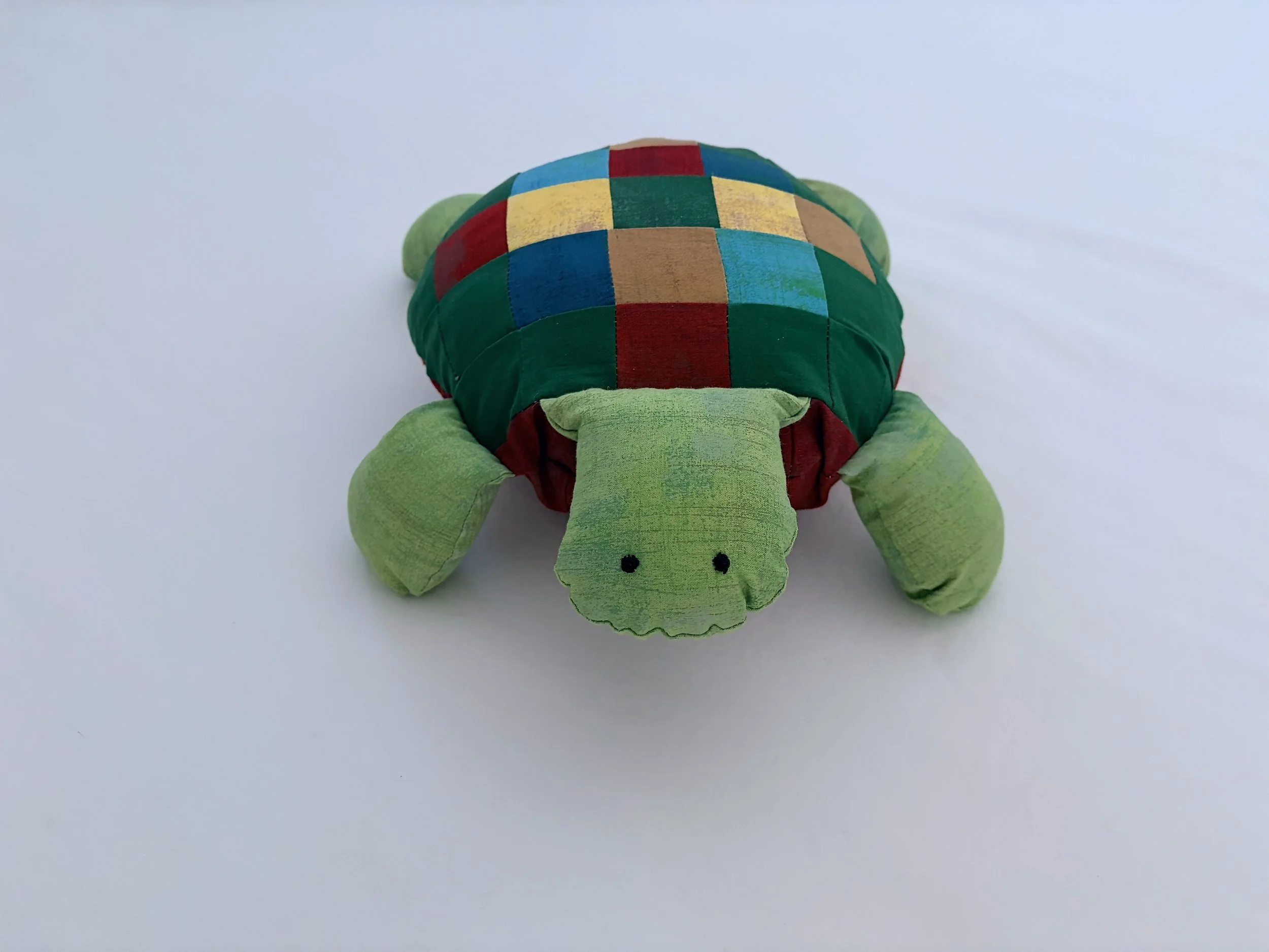 A plush turtle toy with a green head, limbs, and tail, and a colorful checkered shell with red, blue, yellow, brown, and green patches, positioned on a white surface.