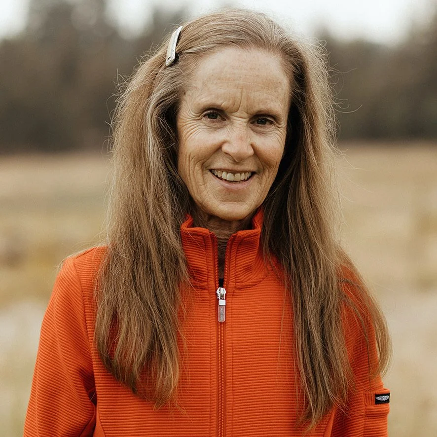 A woman outdoors, wearing an orange zip-up jacket, smiling at the camera, with long brown hair and a hair clip, in a natural landscape background.