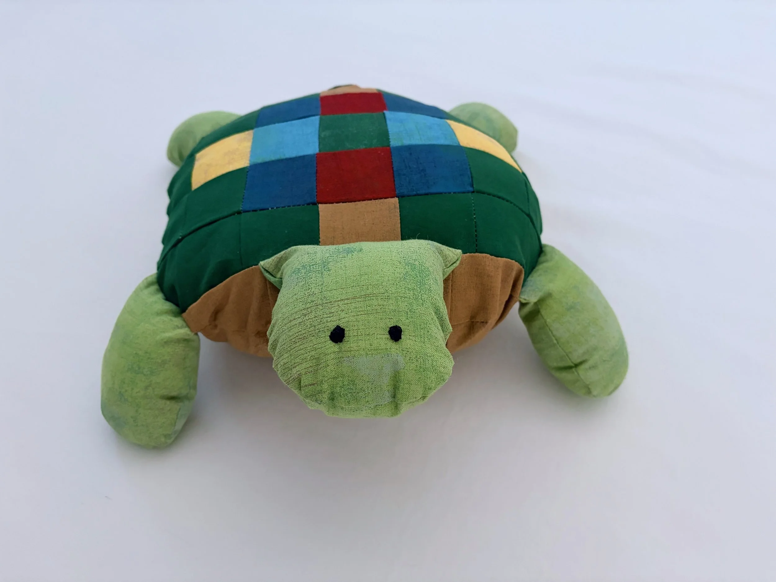 A plush turtle toy with a multicolored checkered shell, green limbs, and a face with black eyes on a white surface.