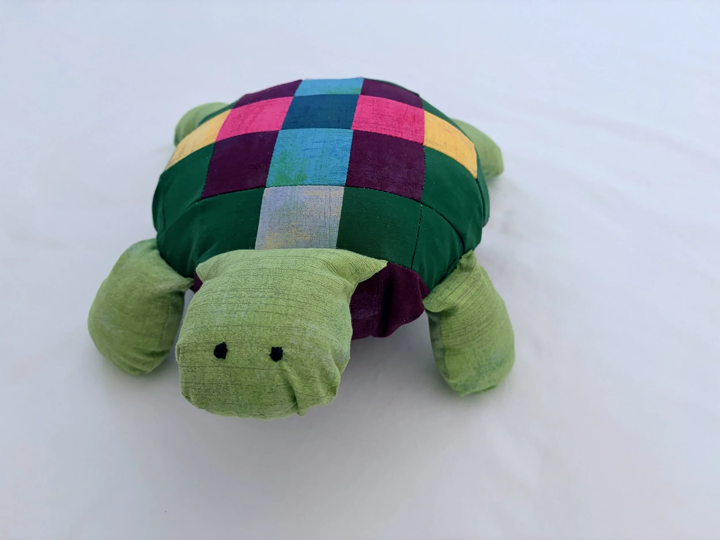 A plush toy turtle with a colorful checkered shell featuring shades of pink, blue, yellow, and purple, set against a plain white background.