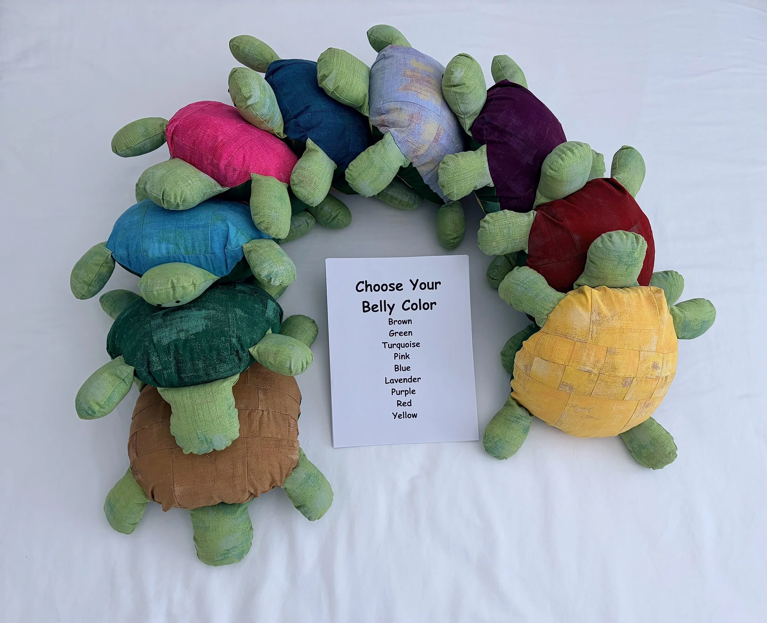Colorful fabric turtles arranged on a white surface with a sign that reads, "Choose Your Belly Color" listing various colors.